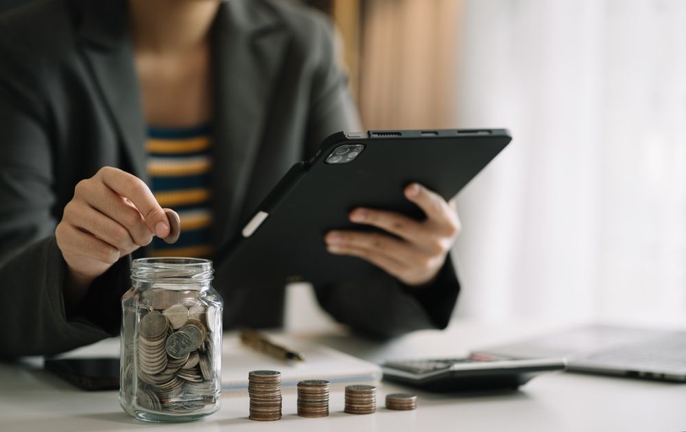 Woman in a suit adds coins to a jar while using a tablet and calculator.