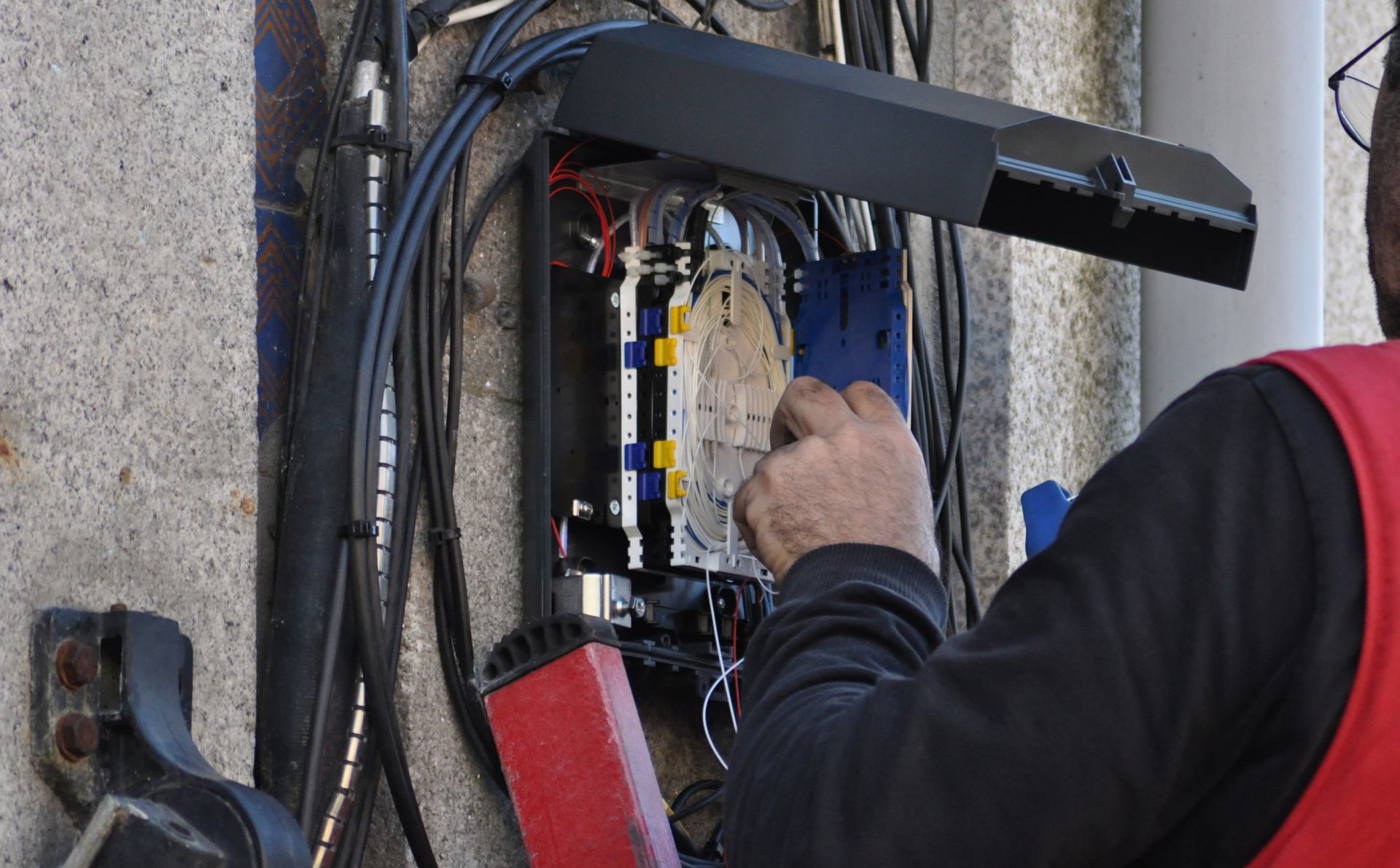 Person working on a network box mounted on a wall. Black cables, blue and yellow components visible. Person working on a network box mounted on a wall. Black cables, blue and yellow components visible.
