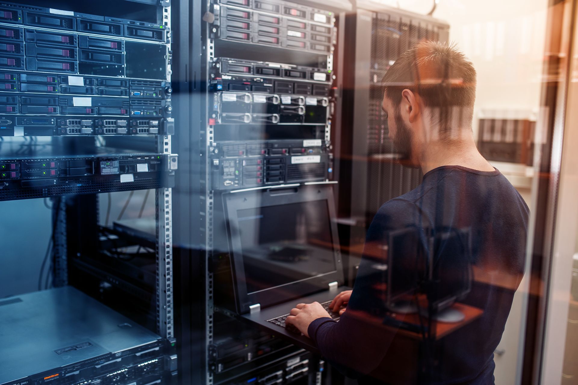 Man working on laptop in a server room with racks of computers.