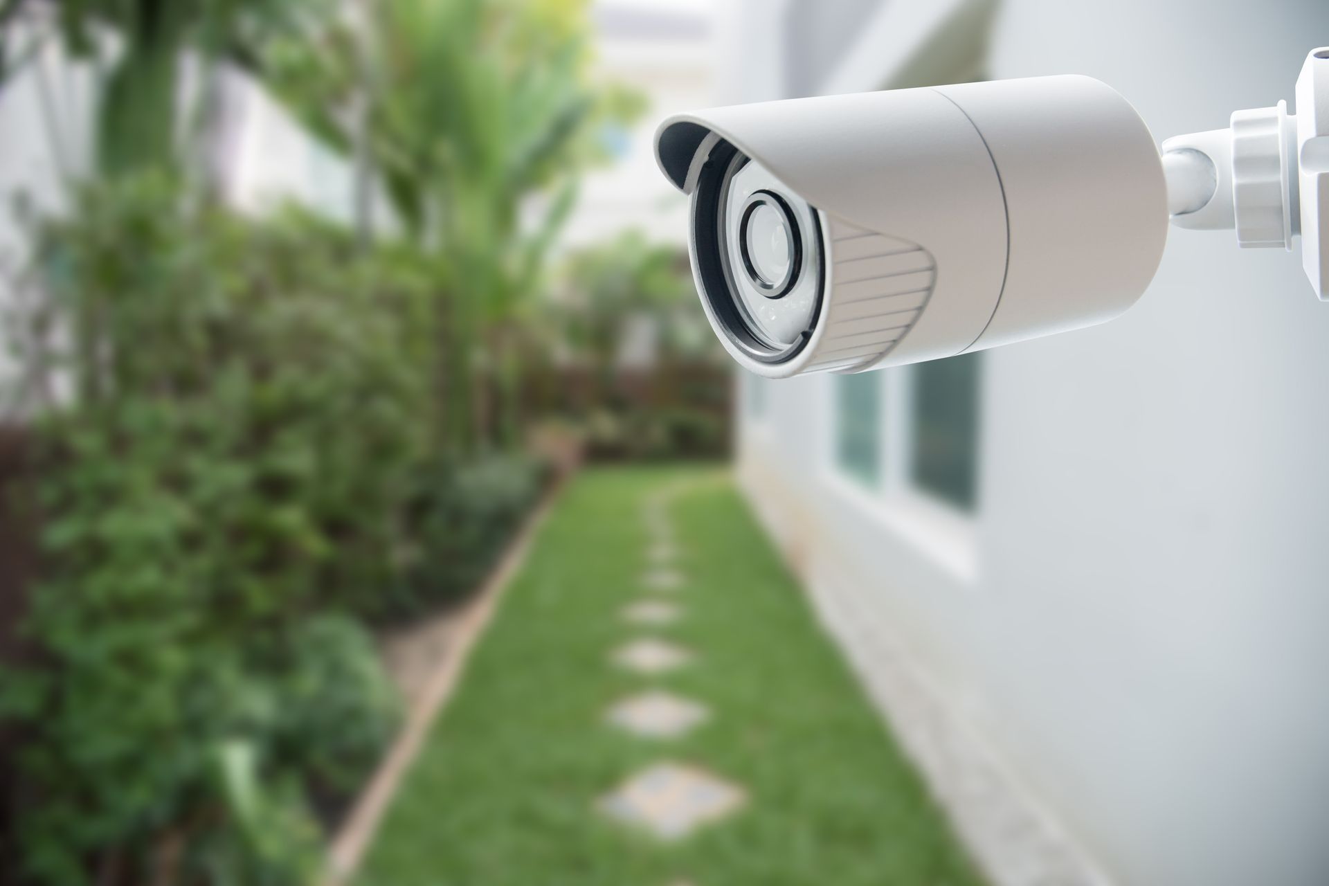 Security camera mounted on a white wall, overlooking a green yard with a stone path.