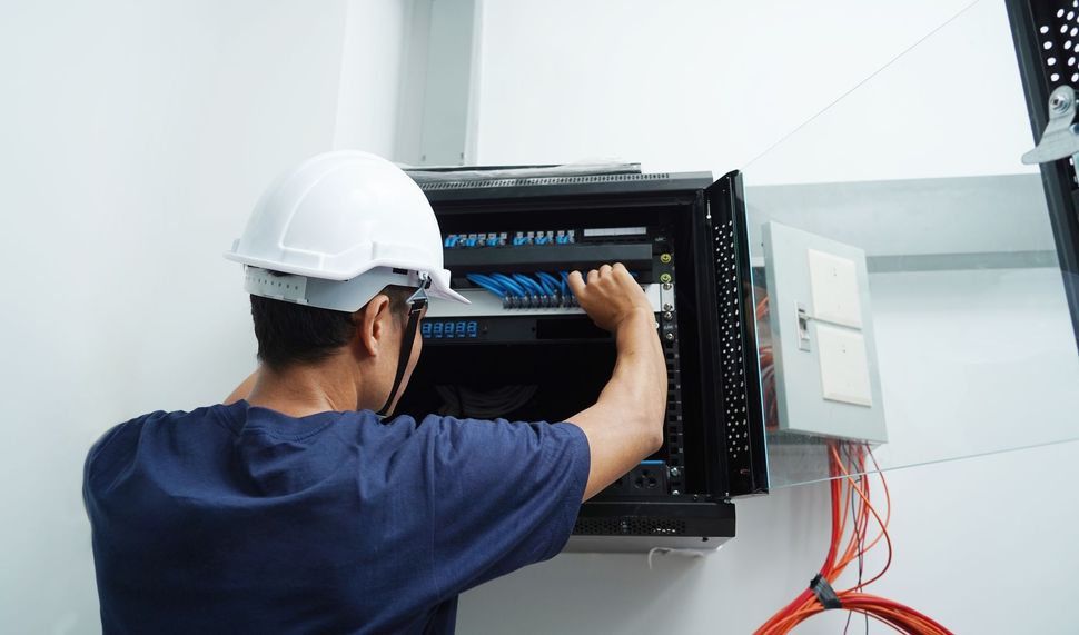 A technician wearing a white hard hat installs network cables into a wall-mounted server rack in a bright room. A technician wearing a white hard hat installs network cables into a wall-mounted server rack in a bright room.