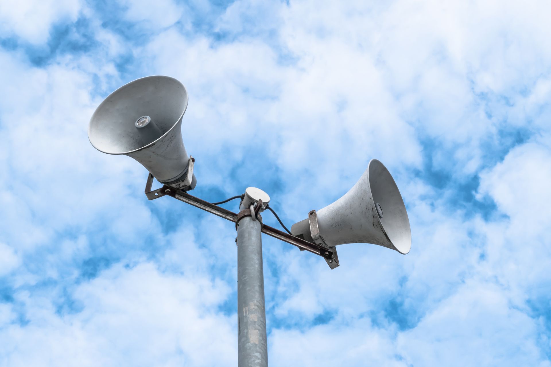 Two gray loudspeakers mounted on a pole against a blue, cloudy sky. Two gray loudspeakers mounted on a pole against a blue, cloudy sky.