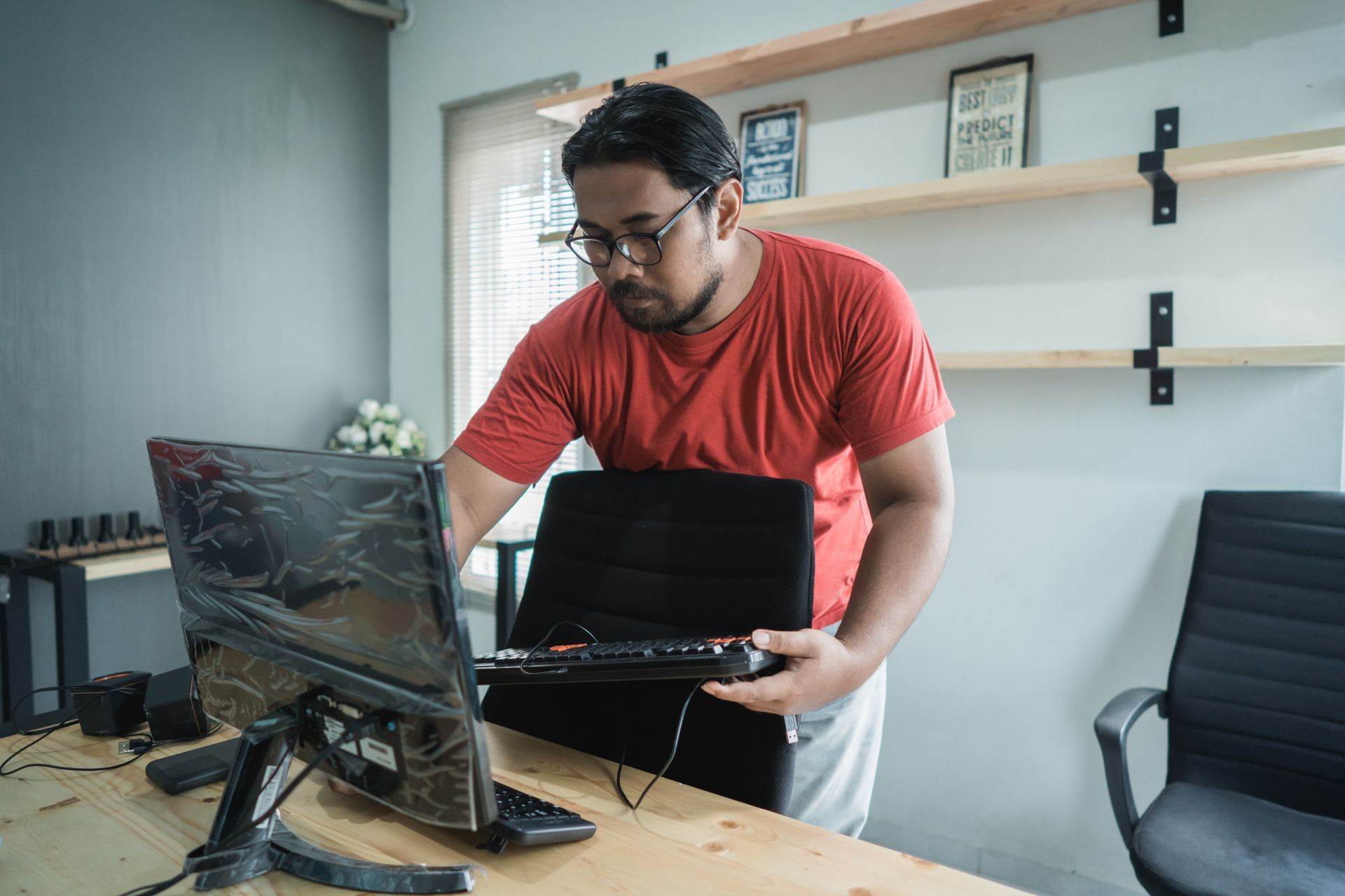 Man wearing glasses, red shirt, working on a computer at a desk in a home office.