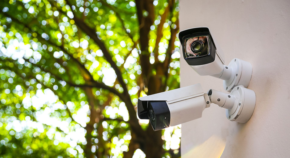Two white security cameras mounted on a light-colored wall against a blurred background of green trees. Two white security cameras mounted on a light-colored wall against a blurred background of green trees.