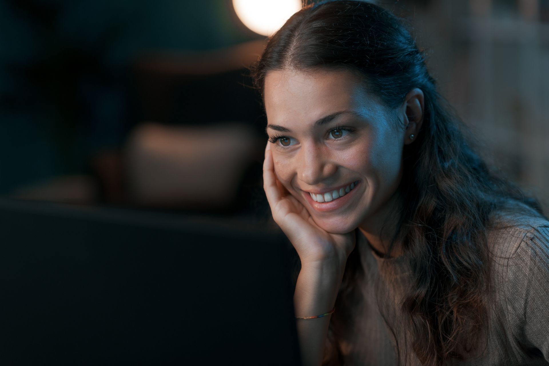 Woman smiling, looking at a laptop screen in a dimly lit room, resting her head on her hand.