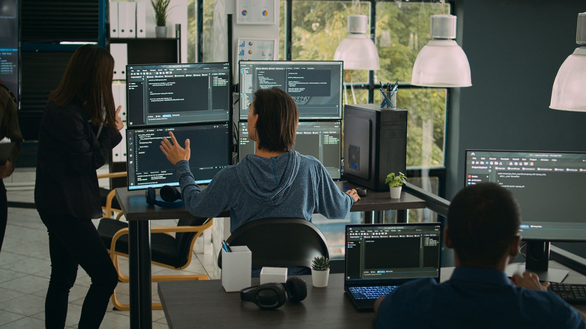 Office scene with people coding at computers; woman talking to a man seated at a desk with multiple screens.