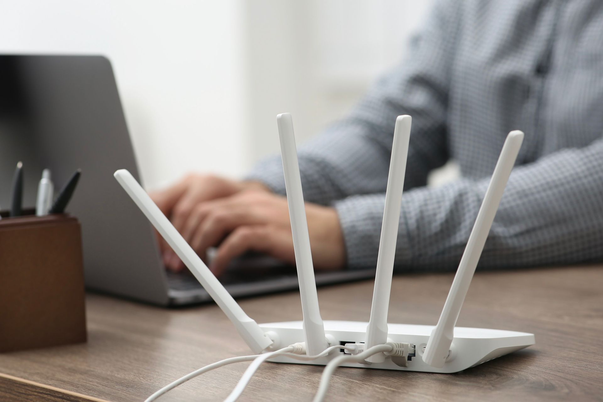 White router with four antennas in front of a person using a laptop on a wooden desk. White router with four antennas in front of a person using a laptop on a wooden desk.