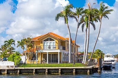 A large house is sitting on a small island in the middle of a body of water surrounded by palm trees.