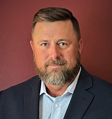 A man with a beard and a suit is standing in front of a red wall.