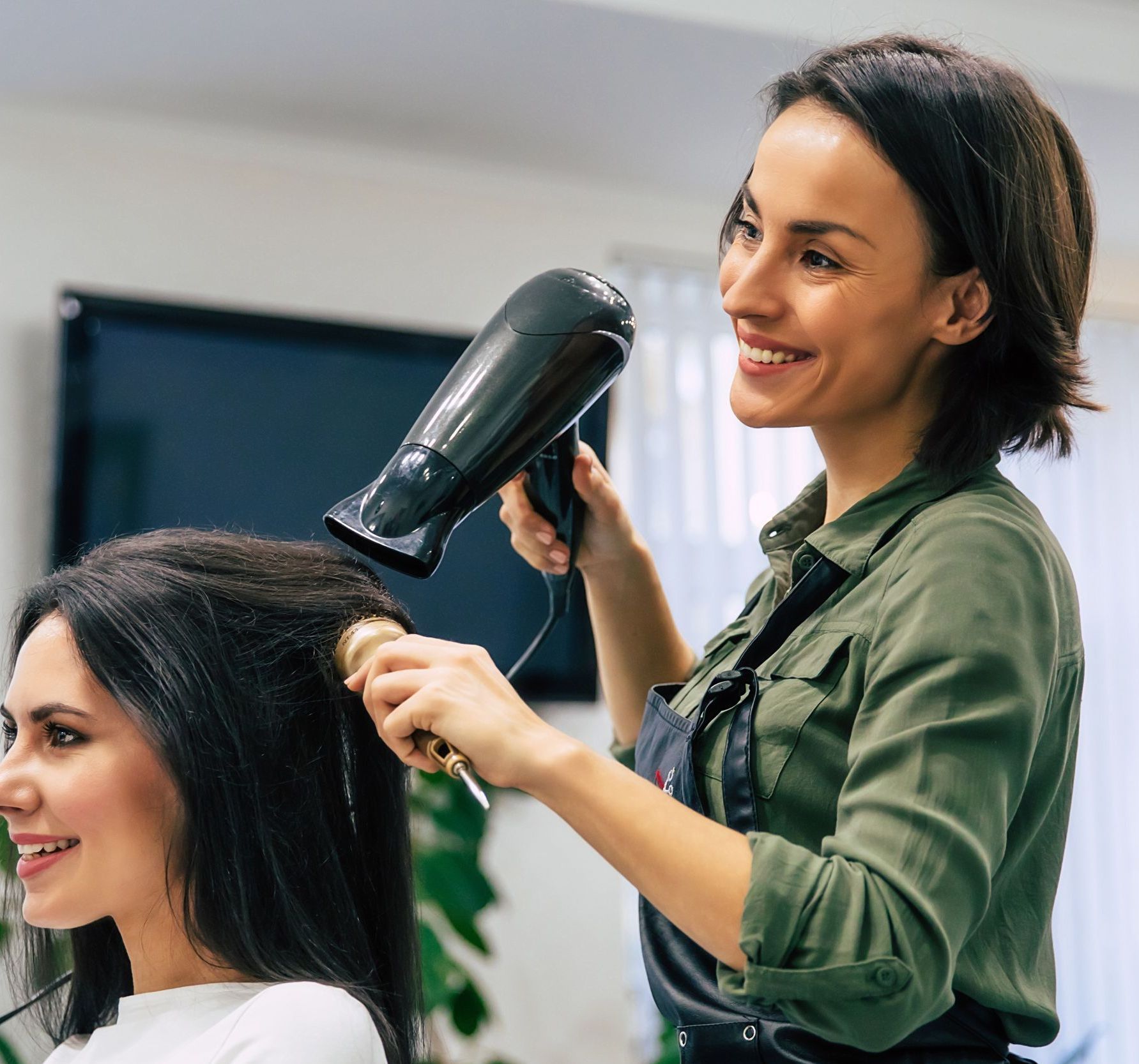 Hair stylist blow-drying client's hair in salon, both smiling. The stylist wears a green shirt and apron.