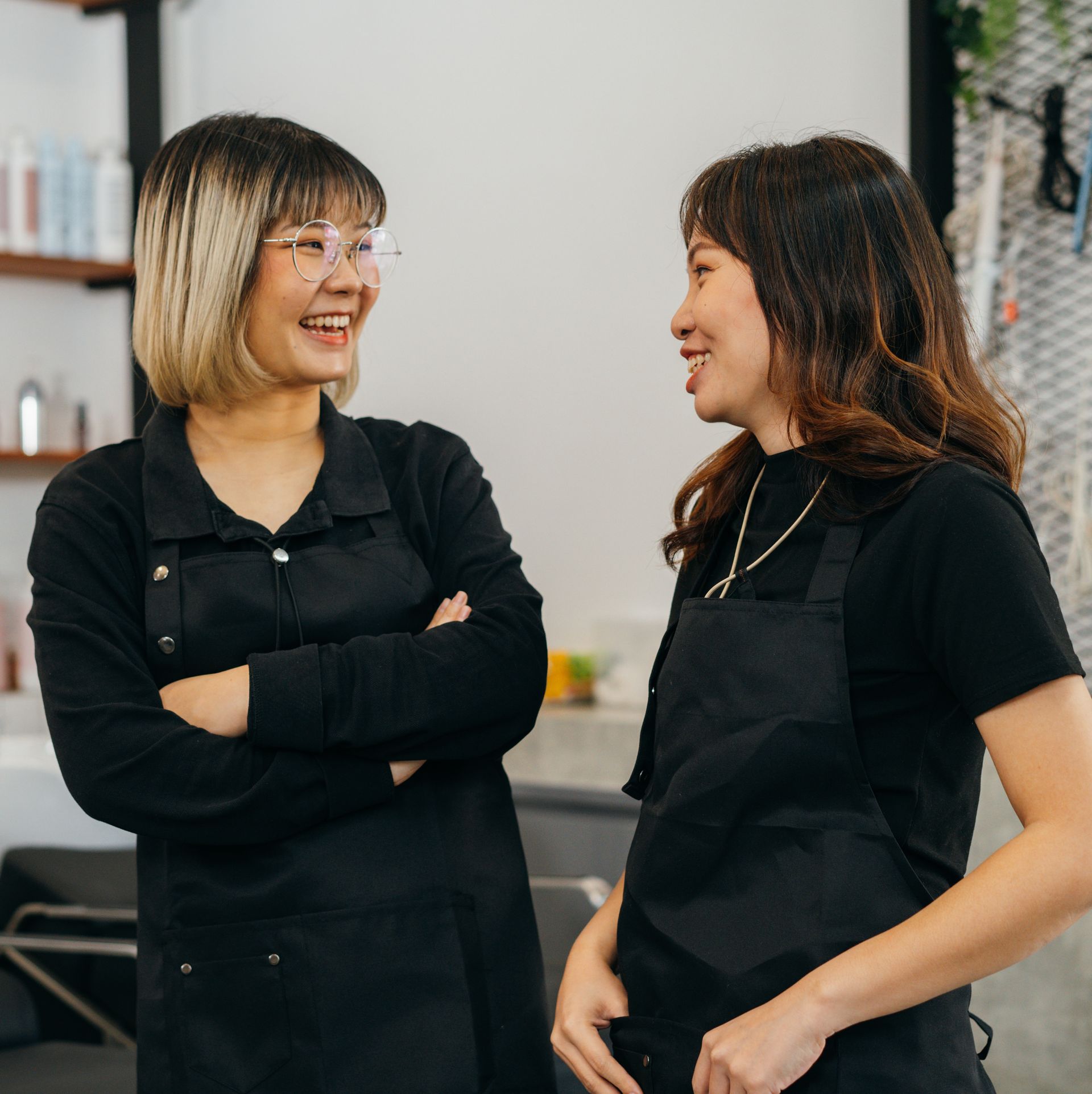 Two women in black aprons laugh while talking in a salon.