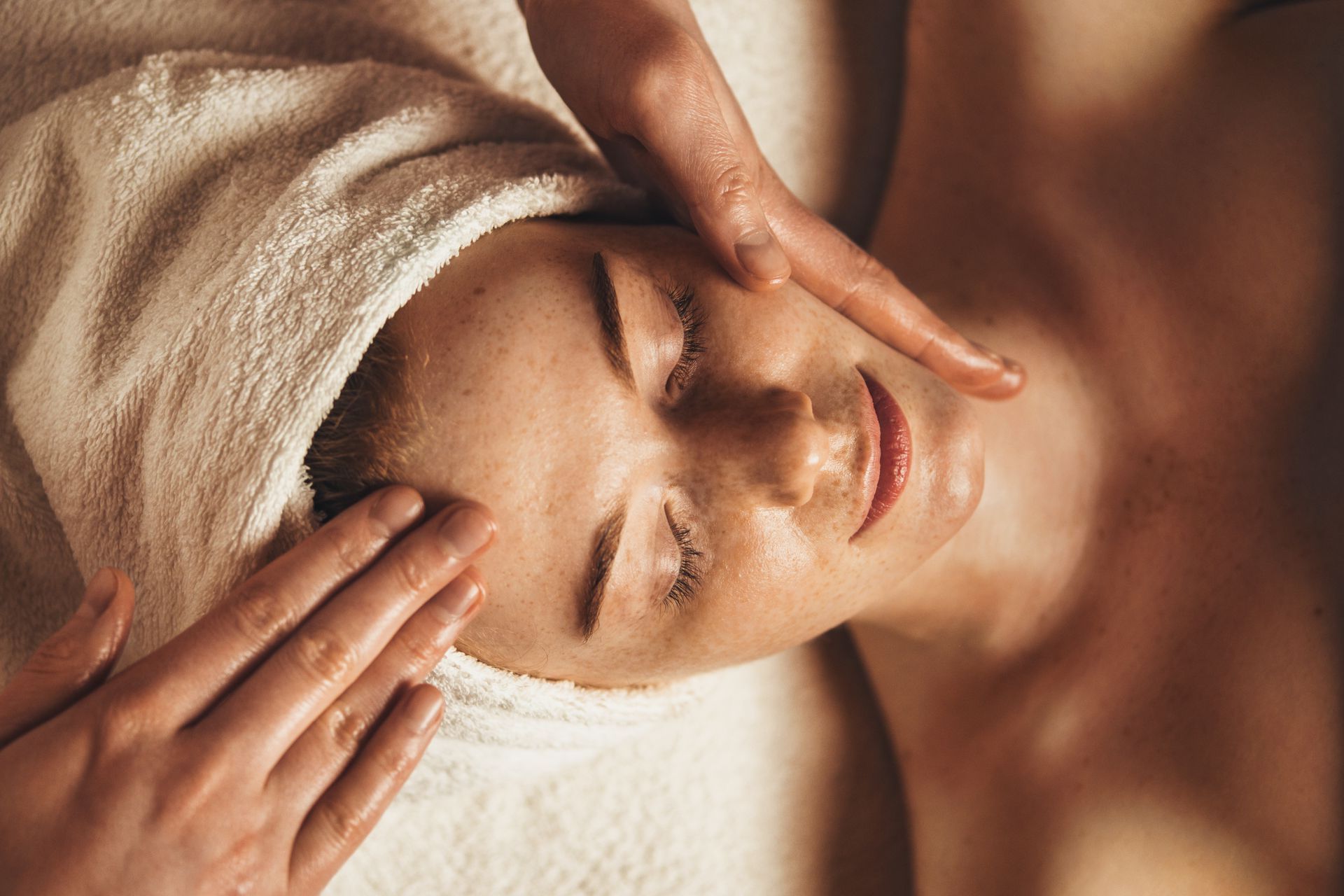 Person receiving a facial massage. Hands on face, eyes closed, towel on head.