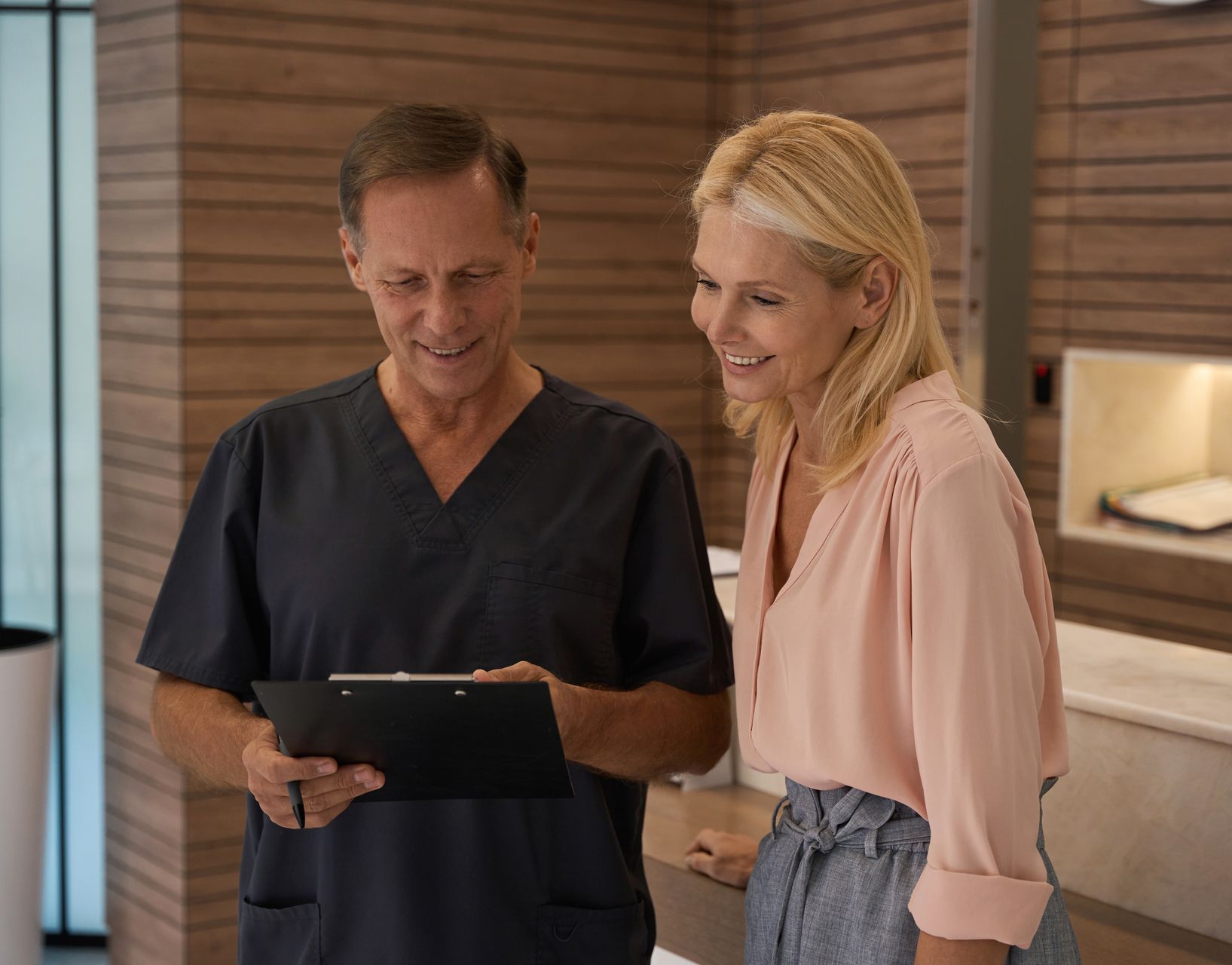A person in scrubs and a woman view a document, smiling, near a desk in a building.