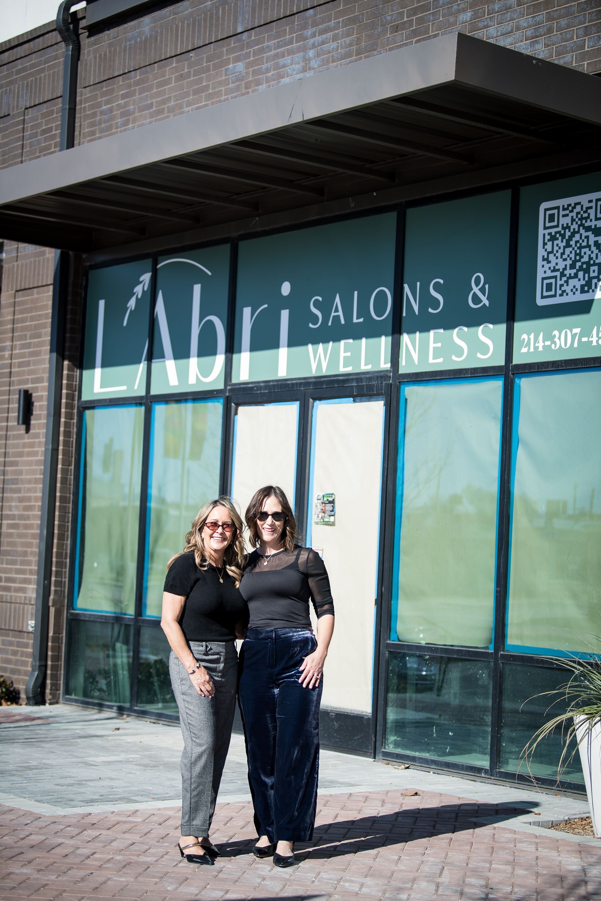 Two women posing together; one wears a black top and blue velvet pants, the other a black top and patterned gray pants.