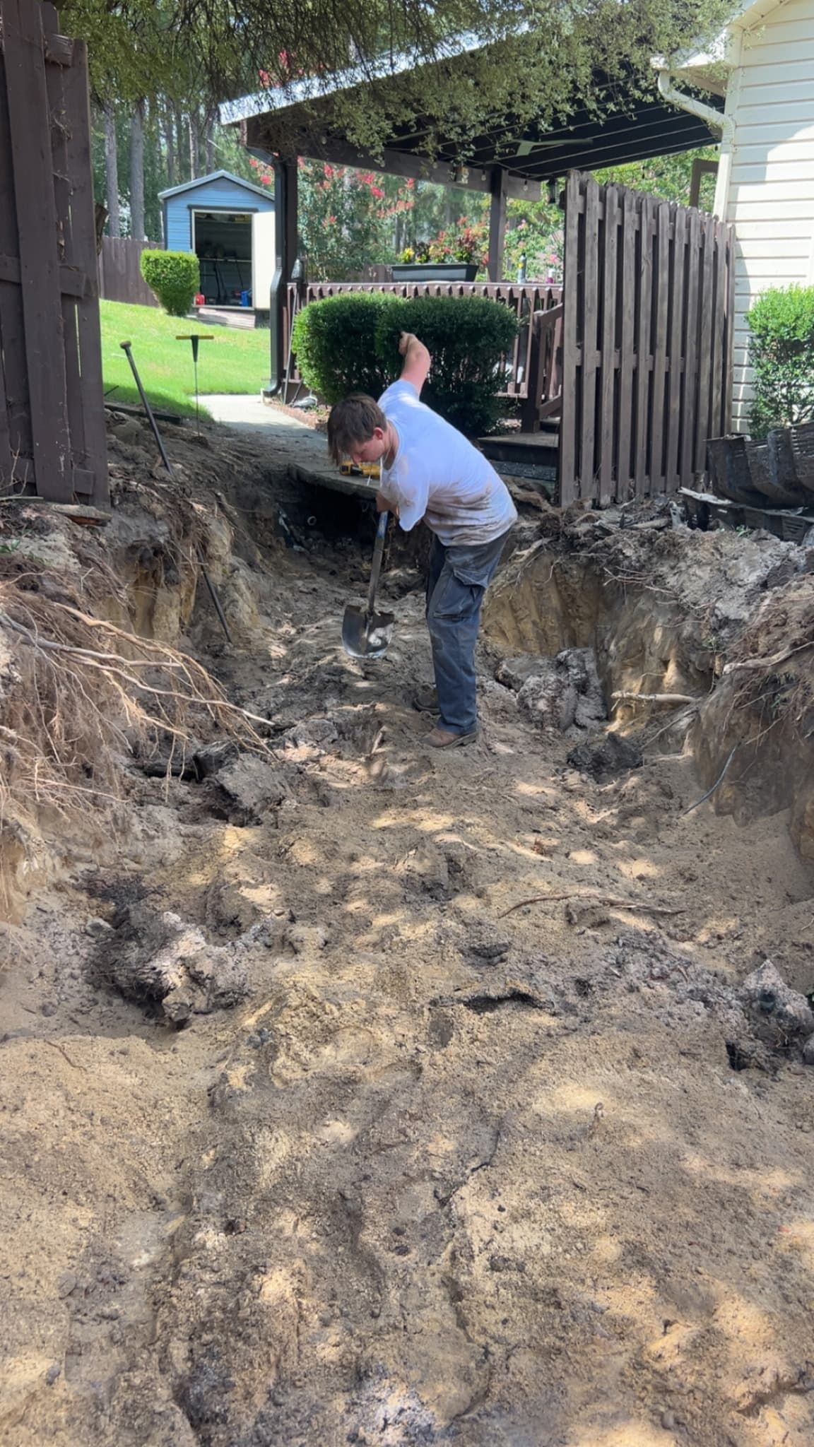 Person digging in a muddy trench beside a house and wooden fence.