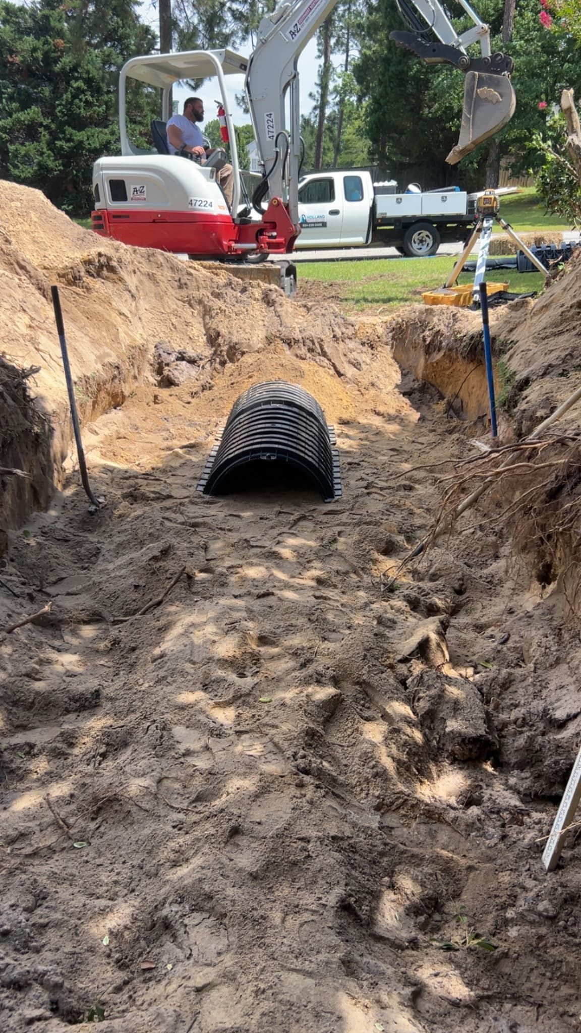 Trenched backyard with a black drainage pipe and excavation equipment on a muddy slope