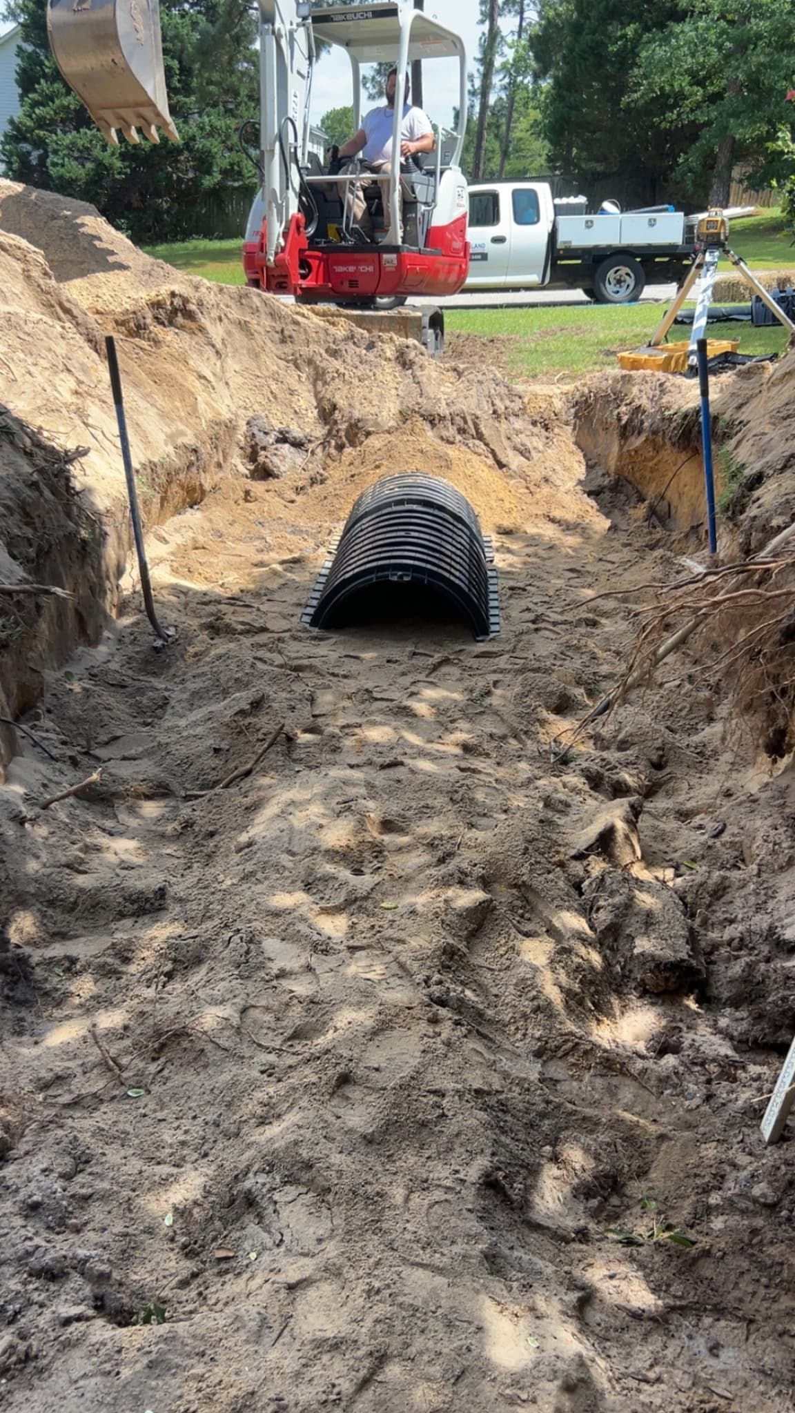 Mud-filled trench with a black drainage pipe, excavator above, and a white utility truck in the background.