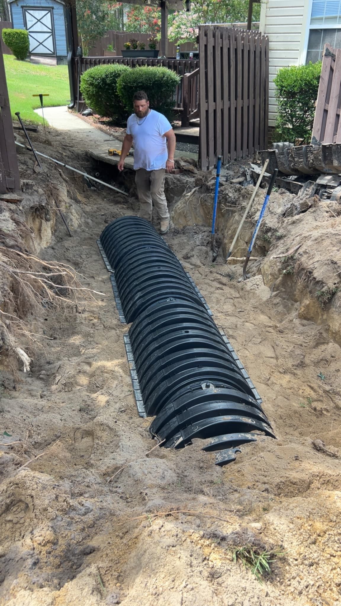 Worker standing beside a large black drainage pipe in a dug trench near a house.
