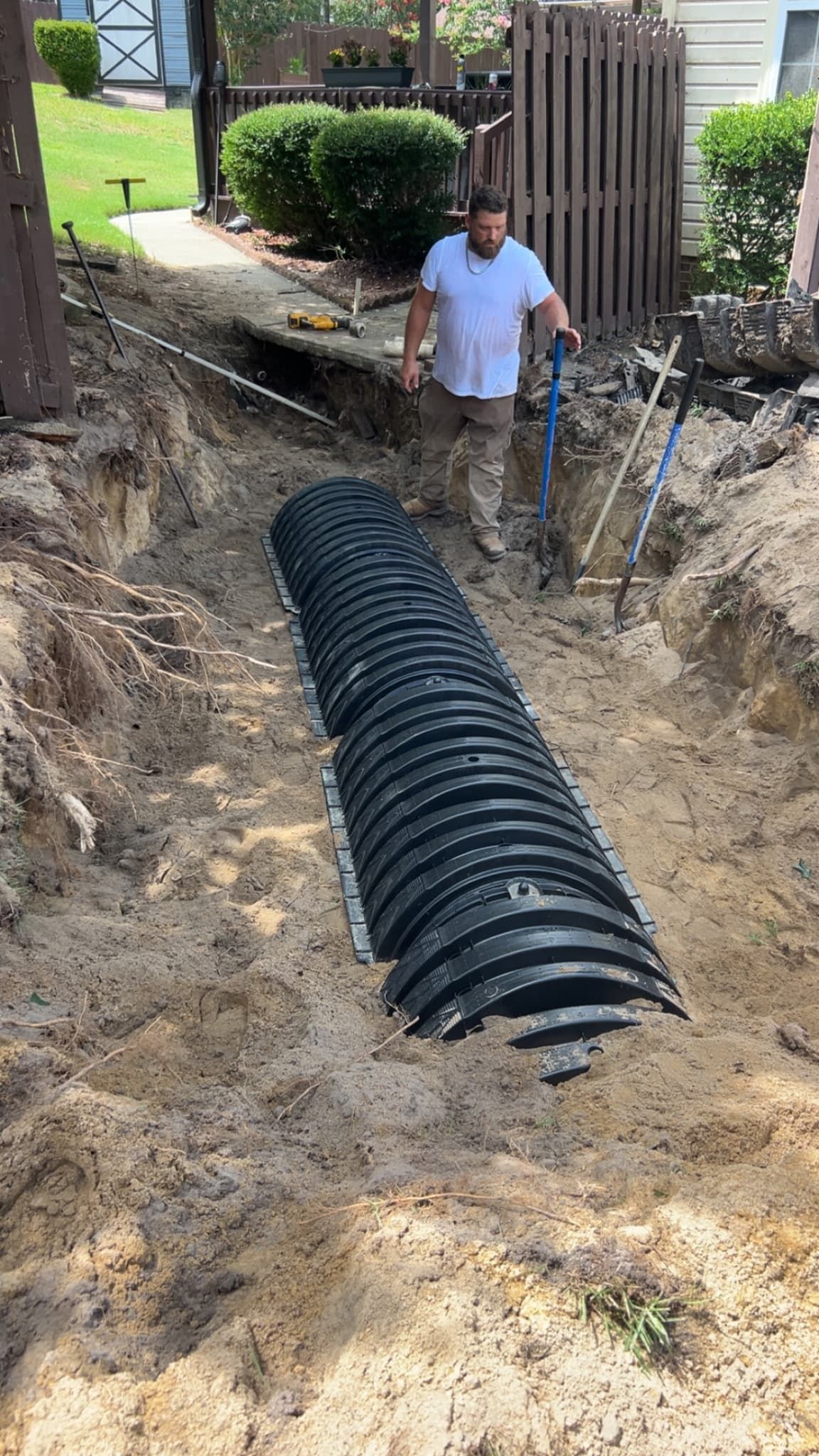 Worker standing in a dug trench beside a black corrugated drainage pipe in a backyard.