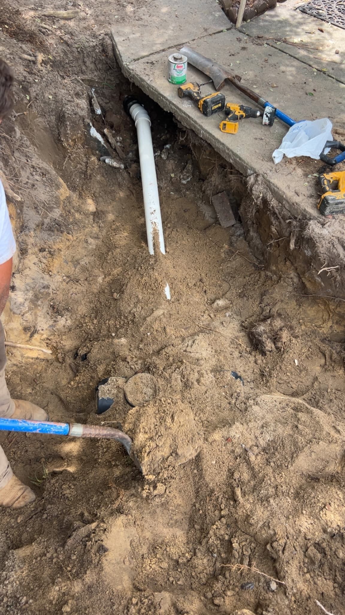Open trench with exposed white pipe, tools on the edge, and muddy soil at a construction site