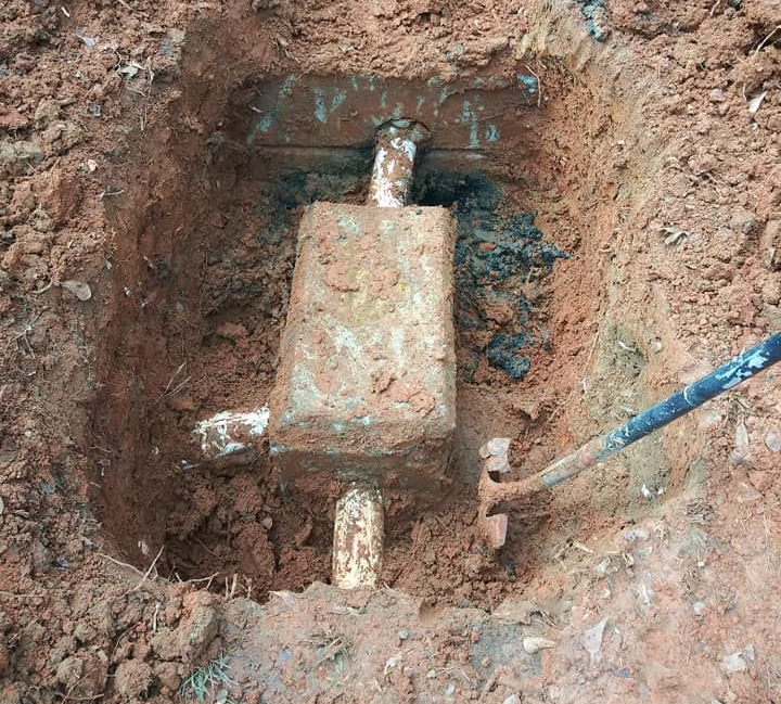 A buried metal valve box in a dirt trench with pipes and a blue-handled tool nearby