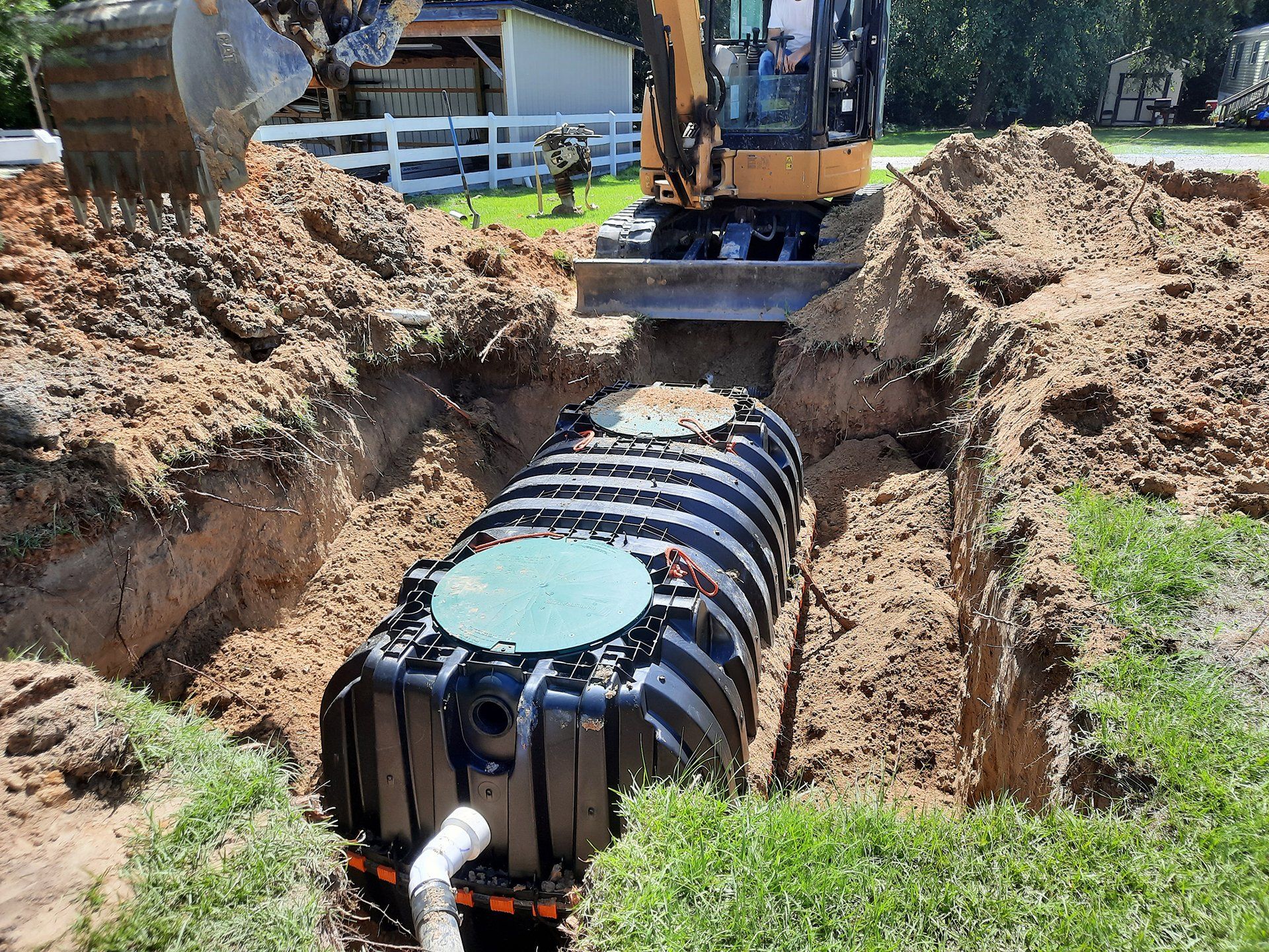 Excavator installing a large black septic tank in a dirt trench at a construction site