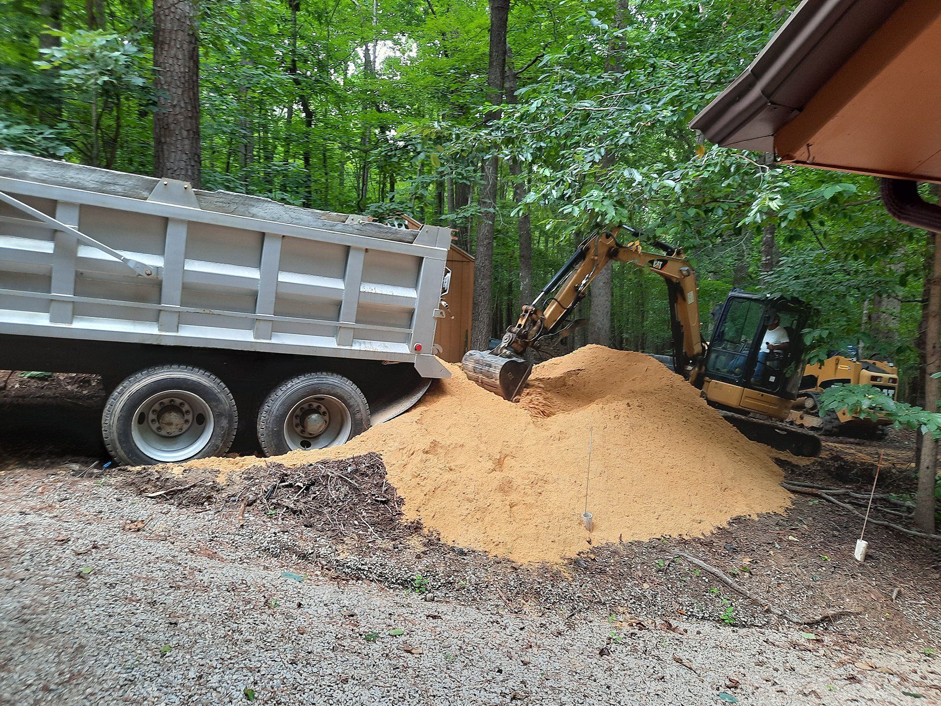 Excavator dumping dirt into a trailer beside a gravel driveway in a wooded area.