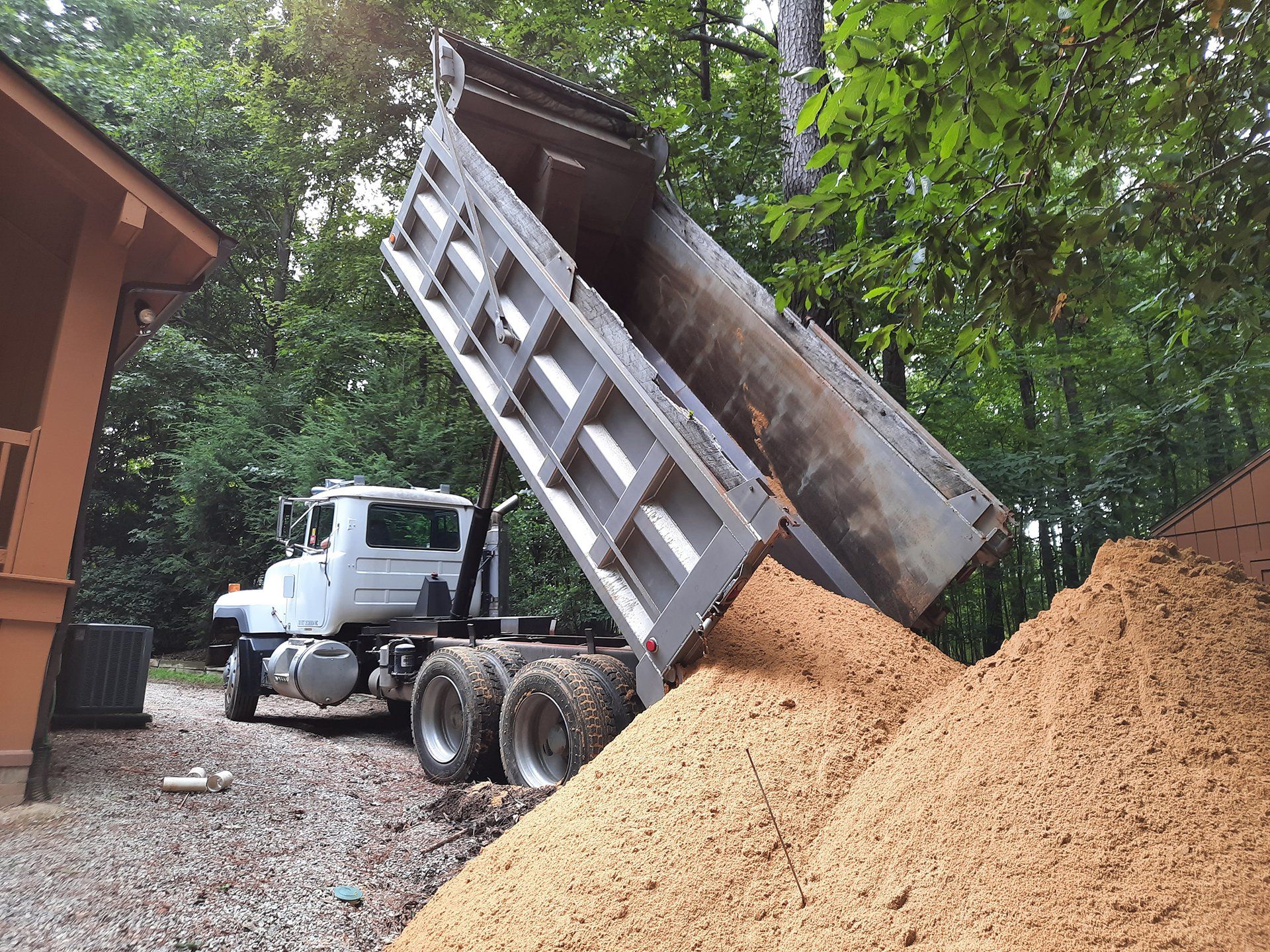 Dump truck unloading a large pile of wood chips beside a building in a wooded area