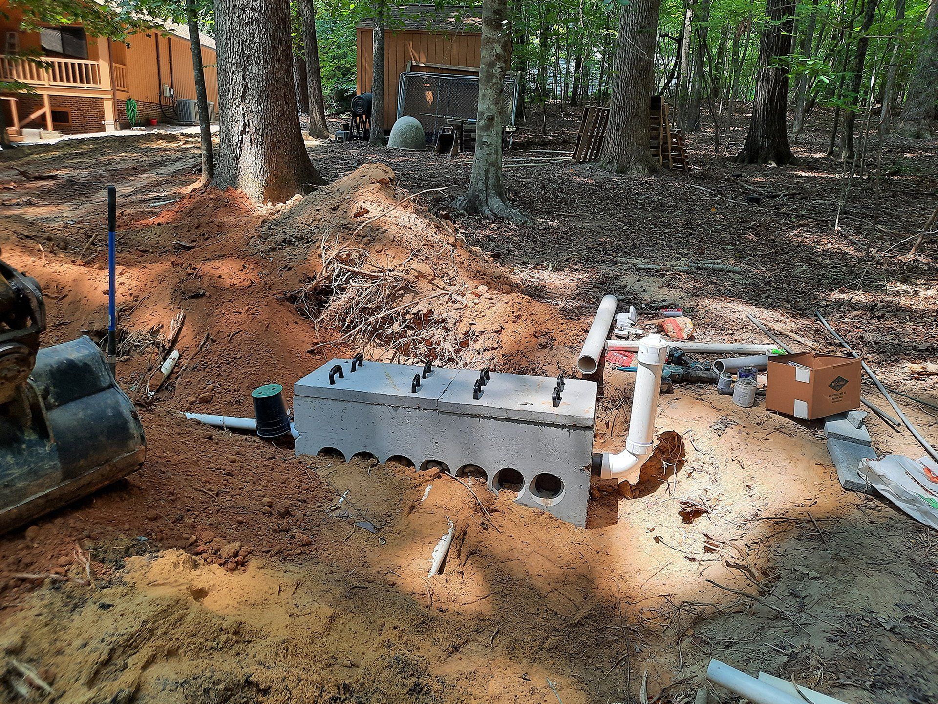 Construction work in a wooded yard with a trench, pipes, soil piles, and equipment exposed