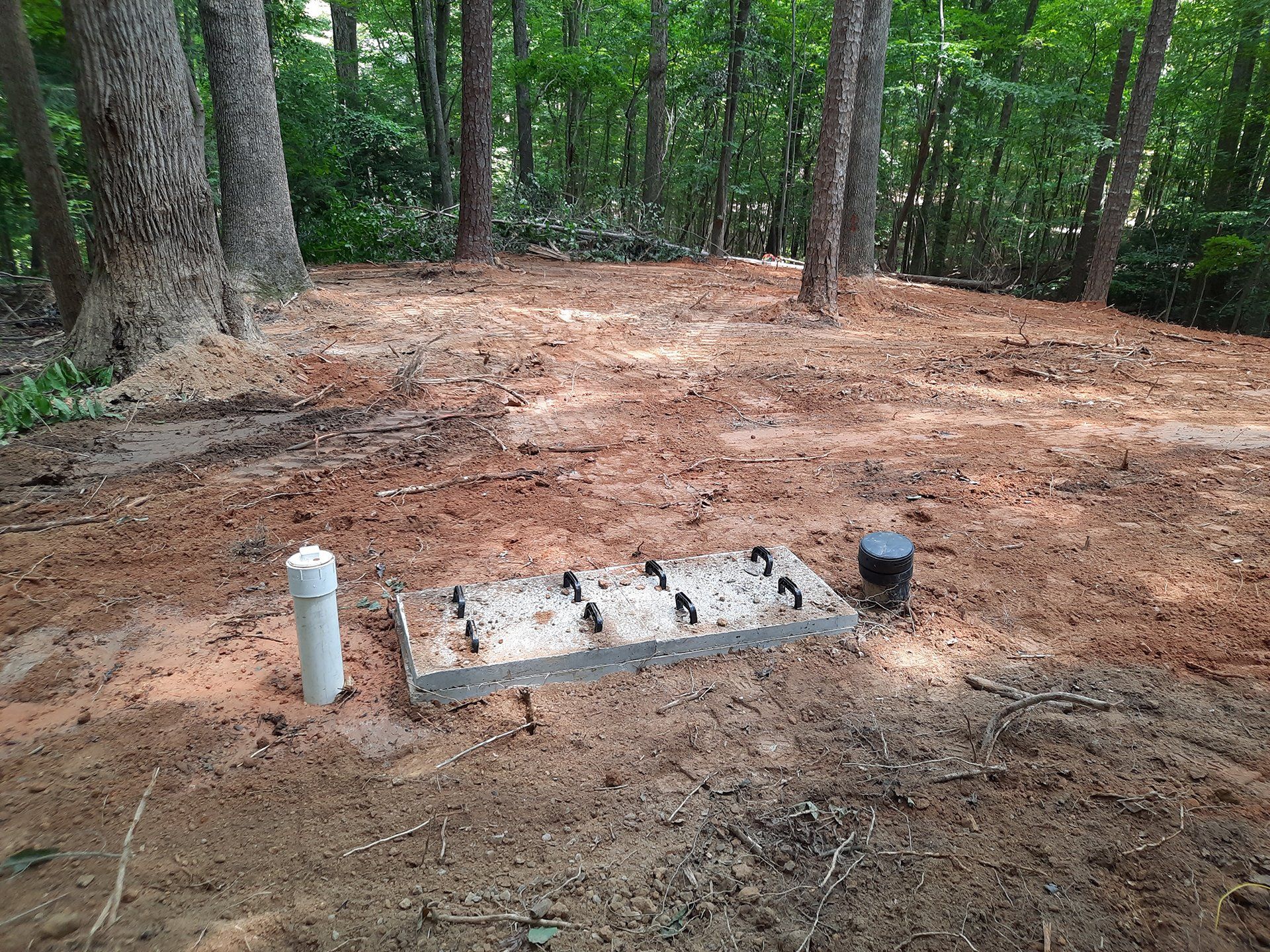 Campsite fire pit with concrete slab and pipes, surrounded by trees and bare ground