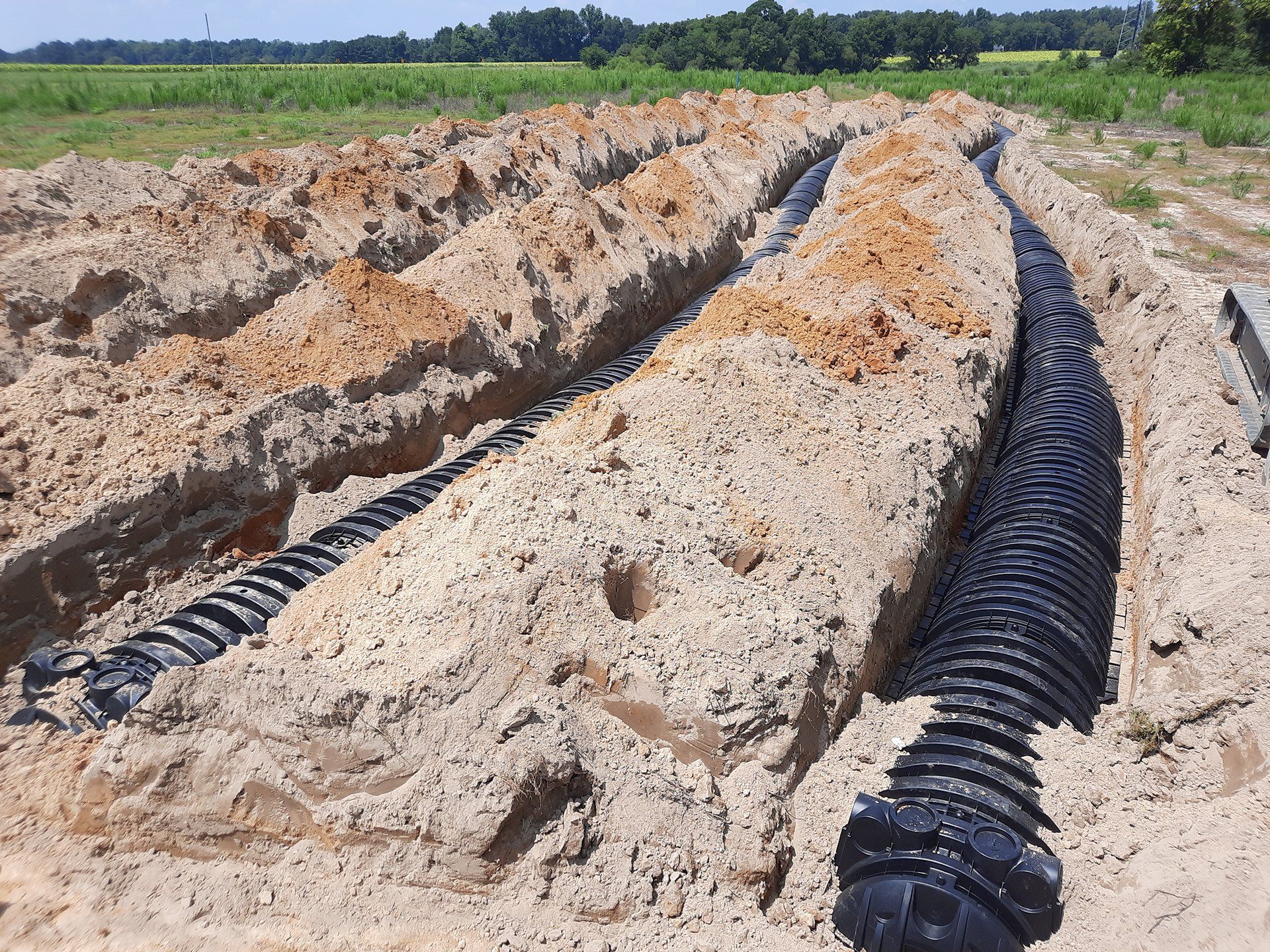 Black drainage pipes laid in trenches across a sandy field under a clear sky