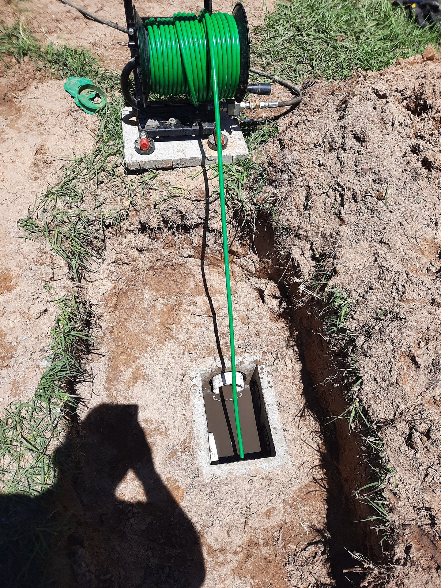 Green cable reel feeding cable into a trench beside an underground box in a dirt yard.