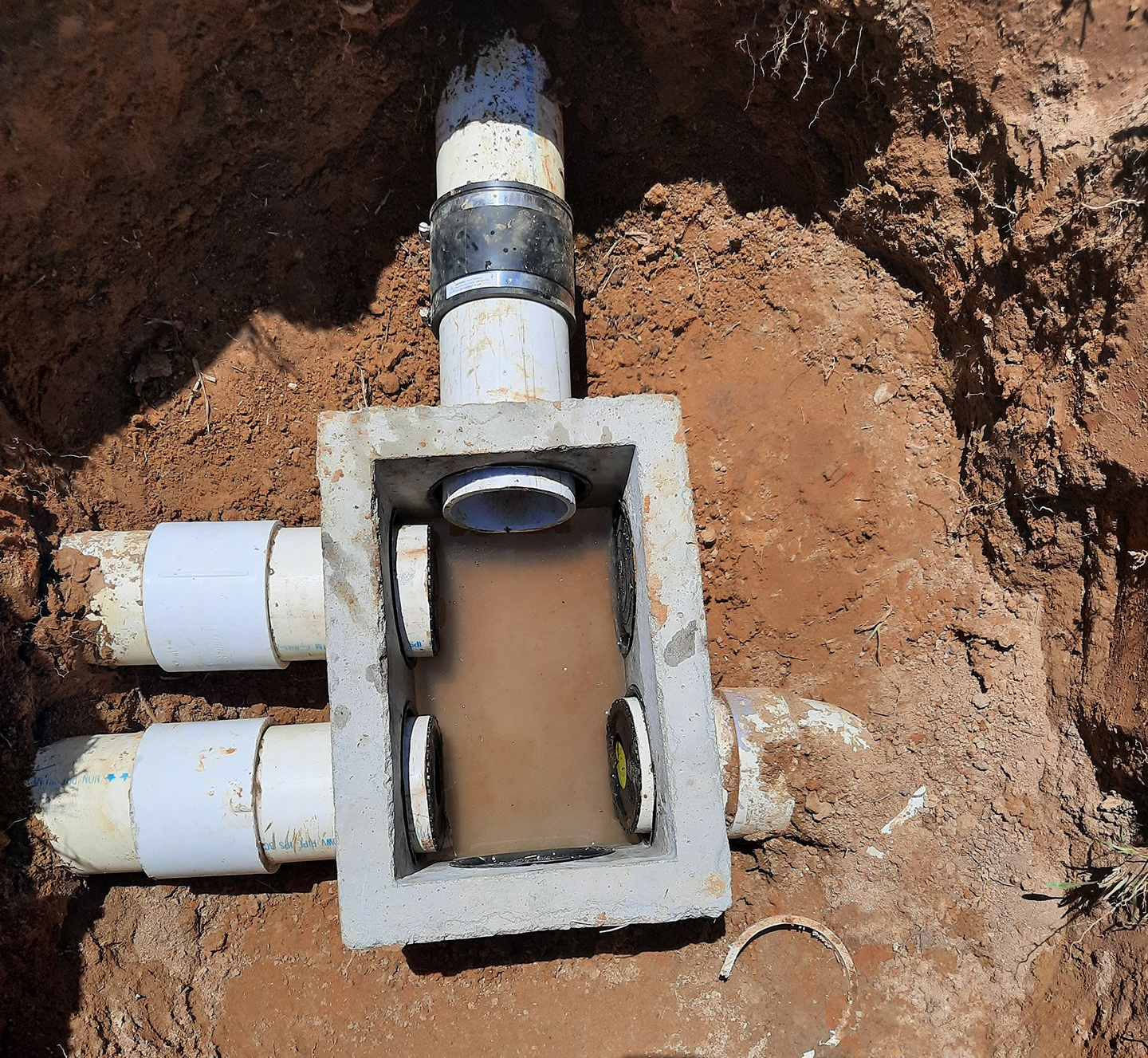 Open utility box in dirt with several white PVC pipes connected to a muddy underground pipe junction