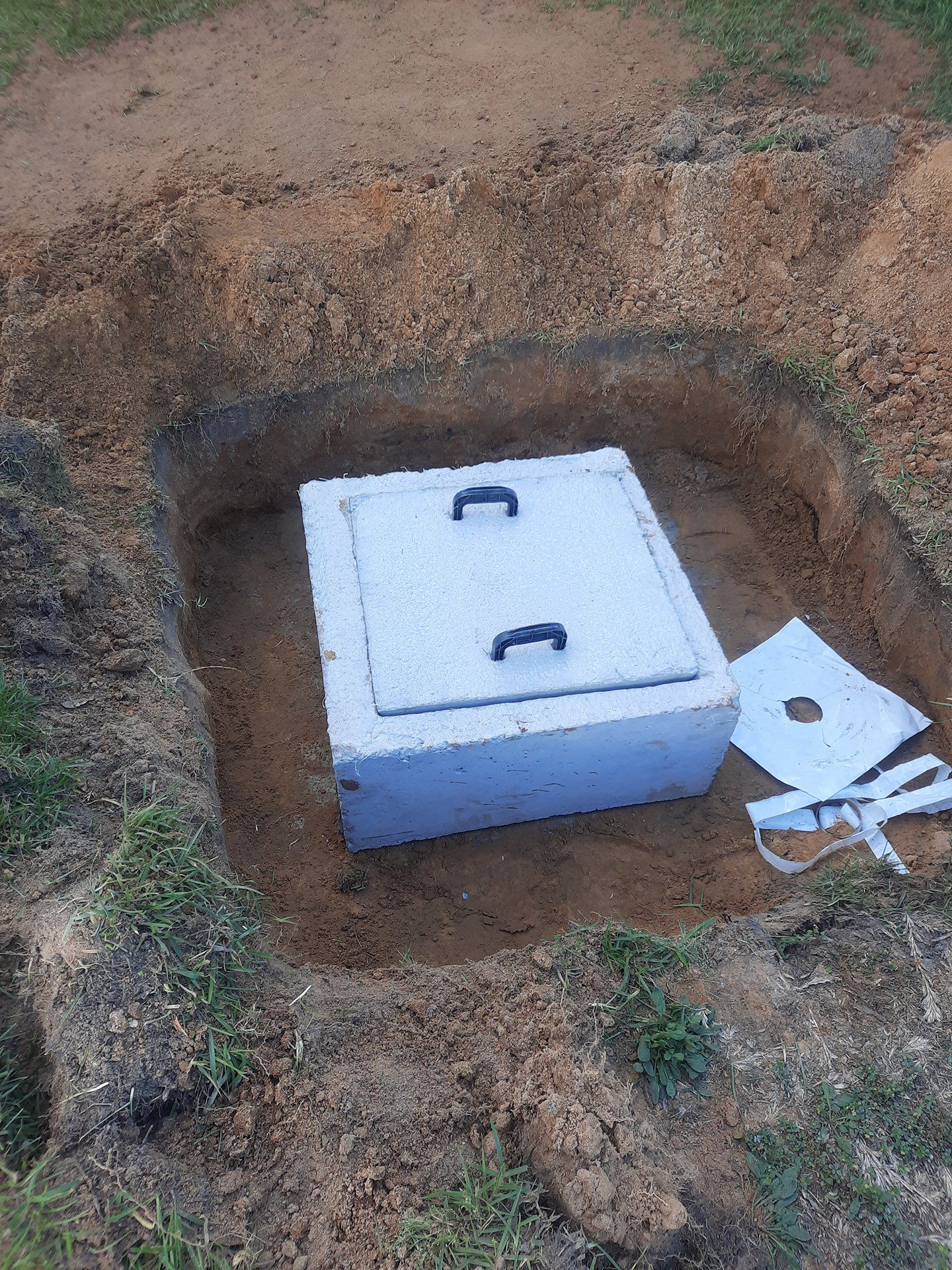 White utility box with handles set in a dirt pit, with a small panel beside it.
