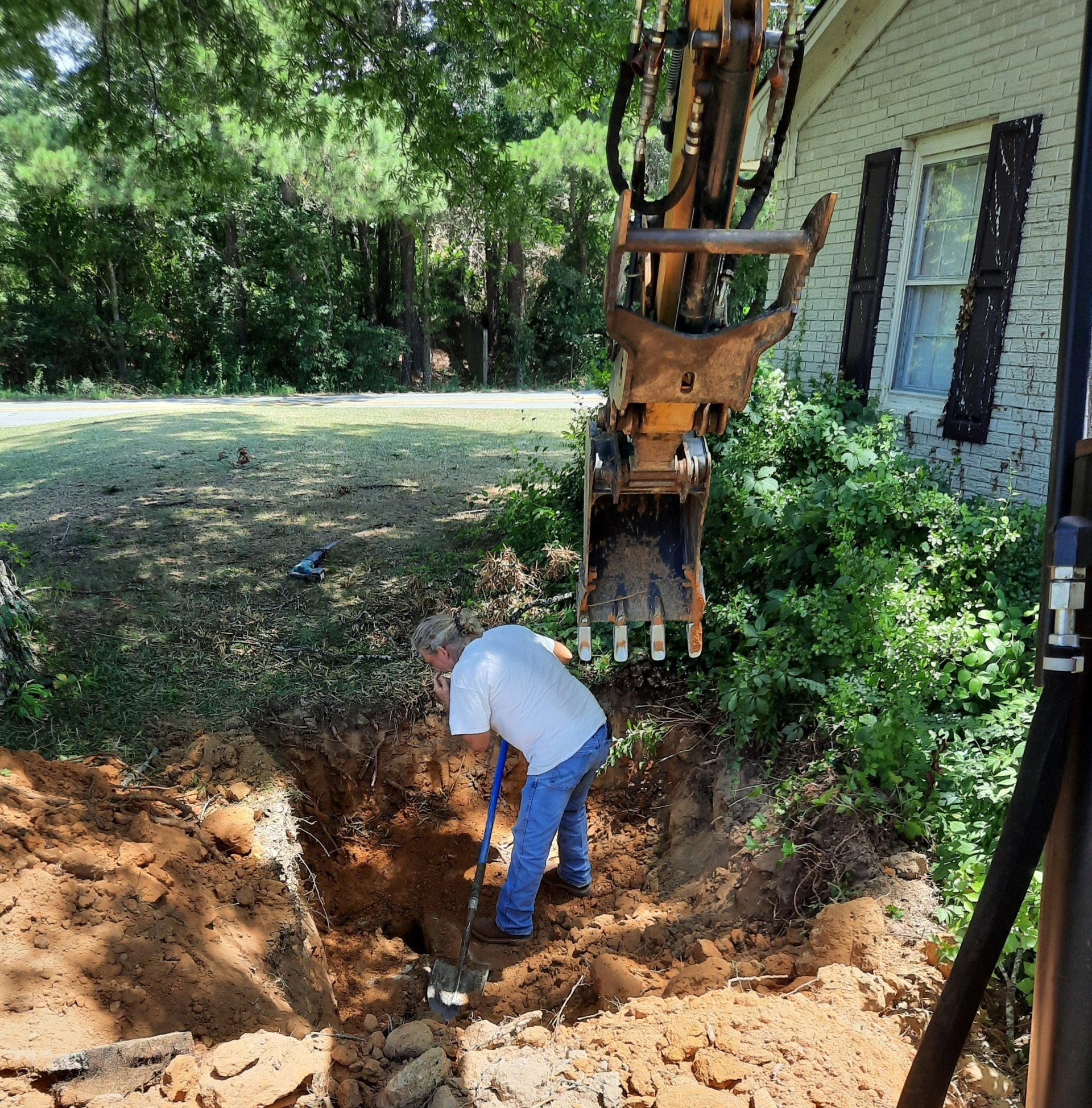 Worker digging a trench beside a house, with an excavator arm overhead and trees in the background