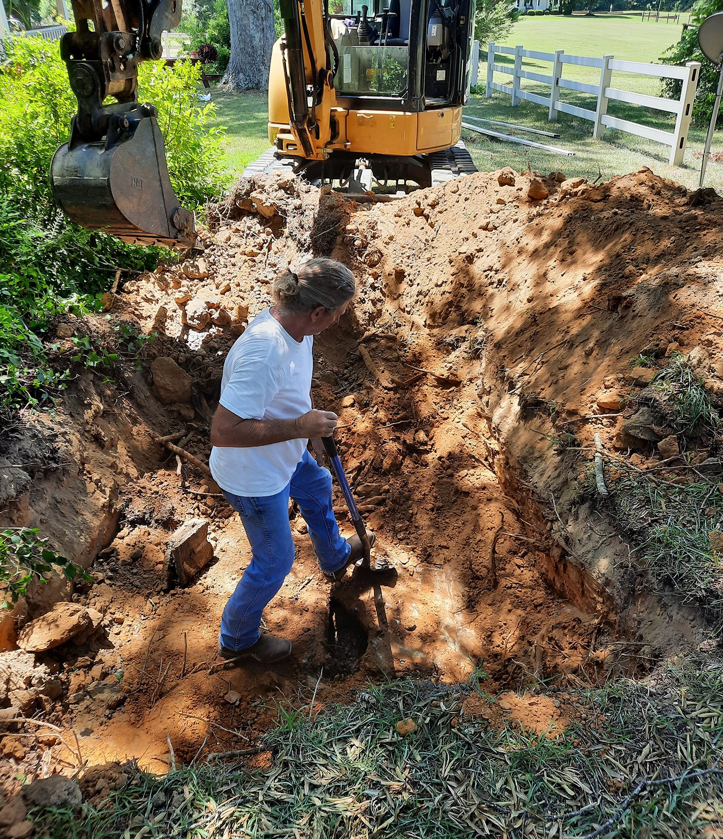 Worker digging soil beside an excavator at a muddy construction site