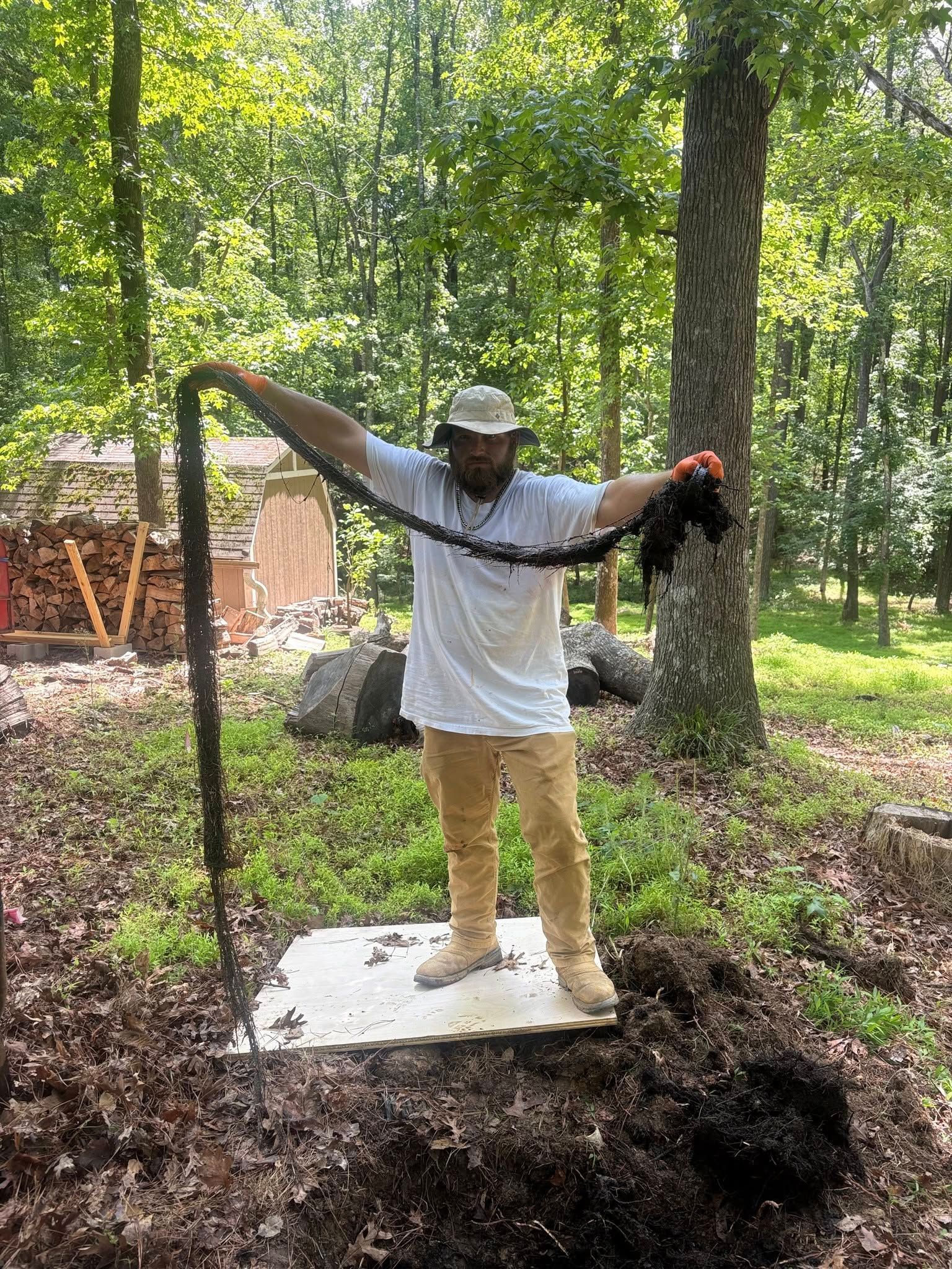 Person standing on a white slab in a wooded area, arms outstretched, holding a long black snake.