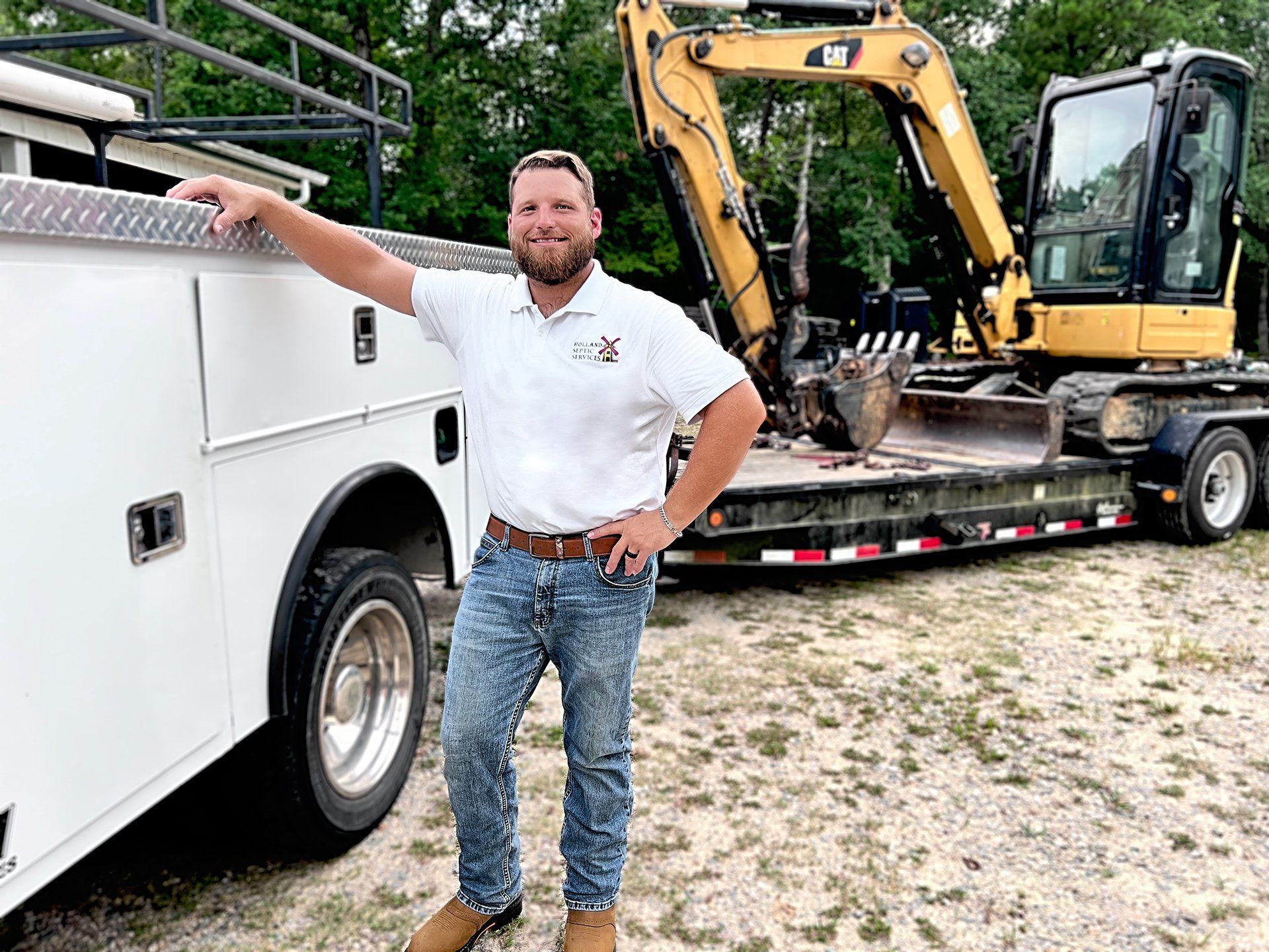 Man standing beside a white service truck and trailer loaded with a yellow excavator outdoors