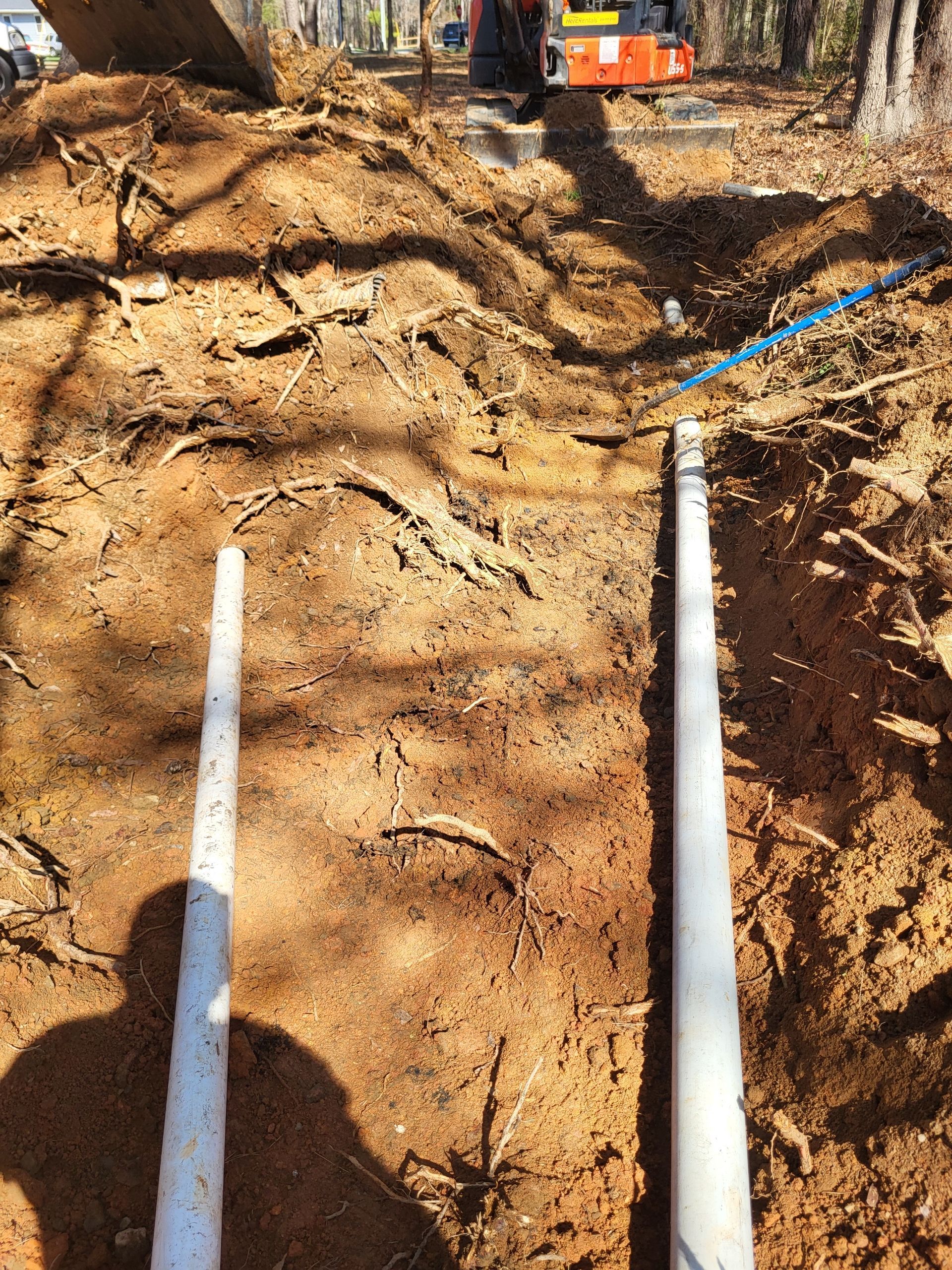 White PVC pipes in a dirt trench at a construction site, with an excavator and equipment in the background