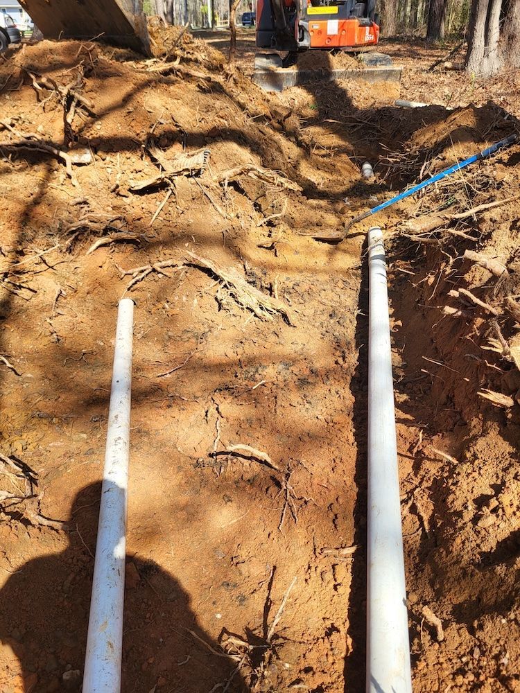 Trench with two white pipes laid in dirt, construction equipment visible in the background.