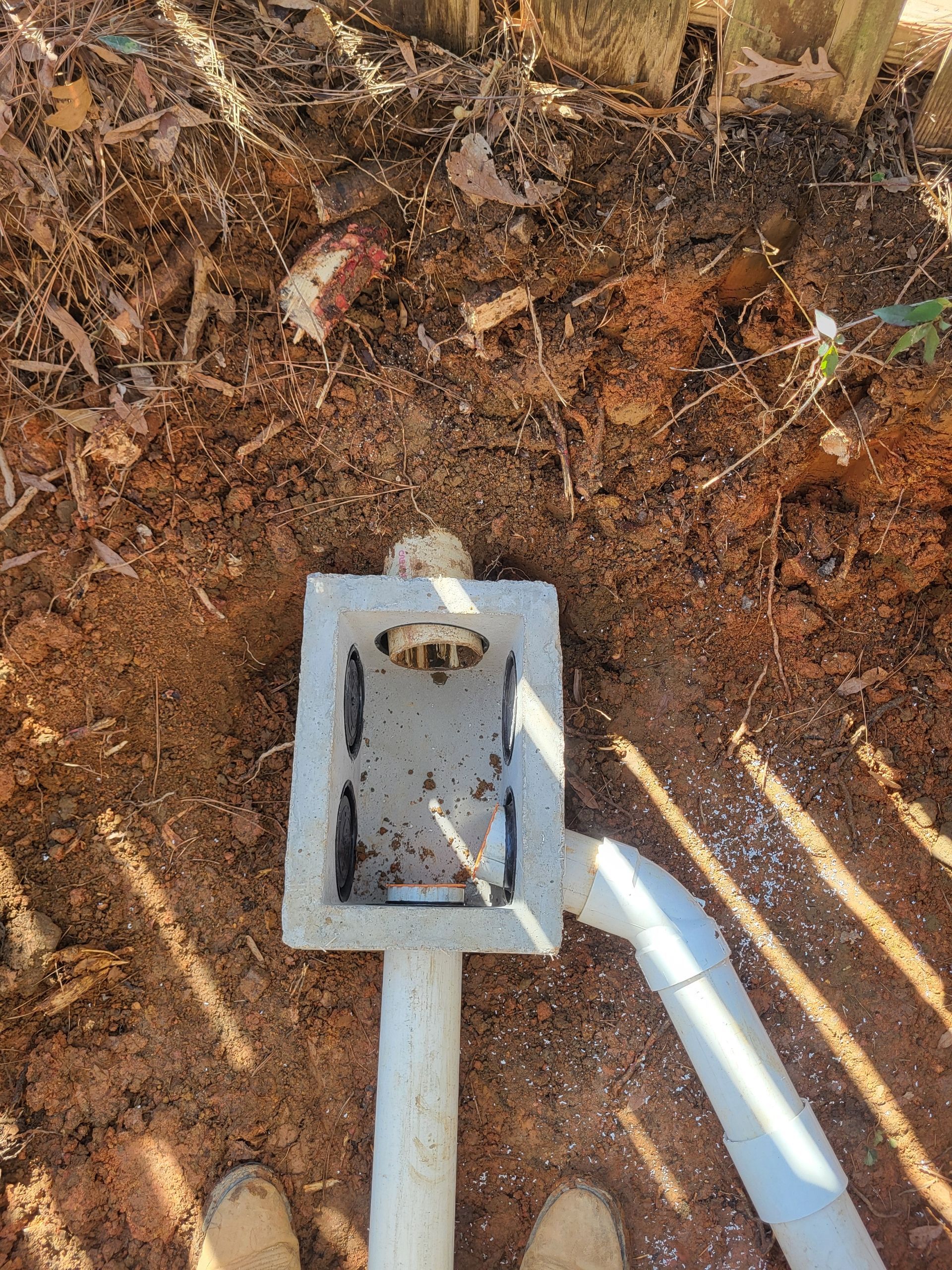 Concrete electrical box in dirt beside a white conduit pipe, with dry leaves and shadows nearby