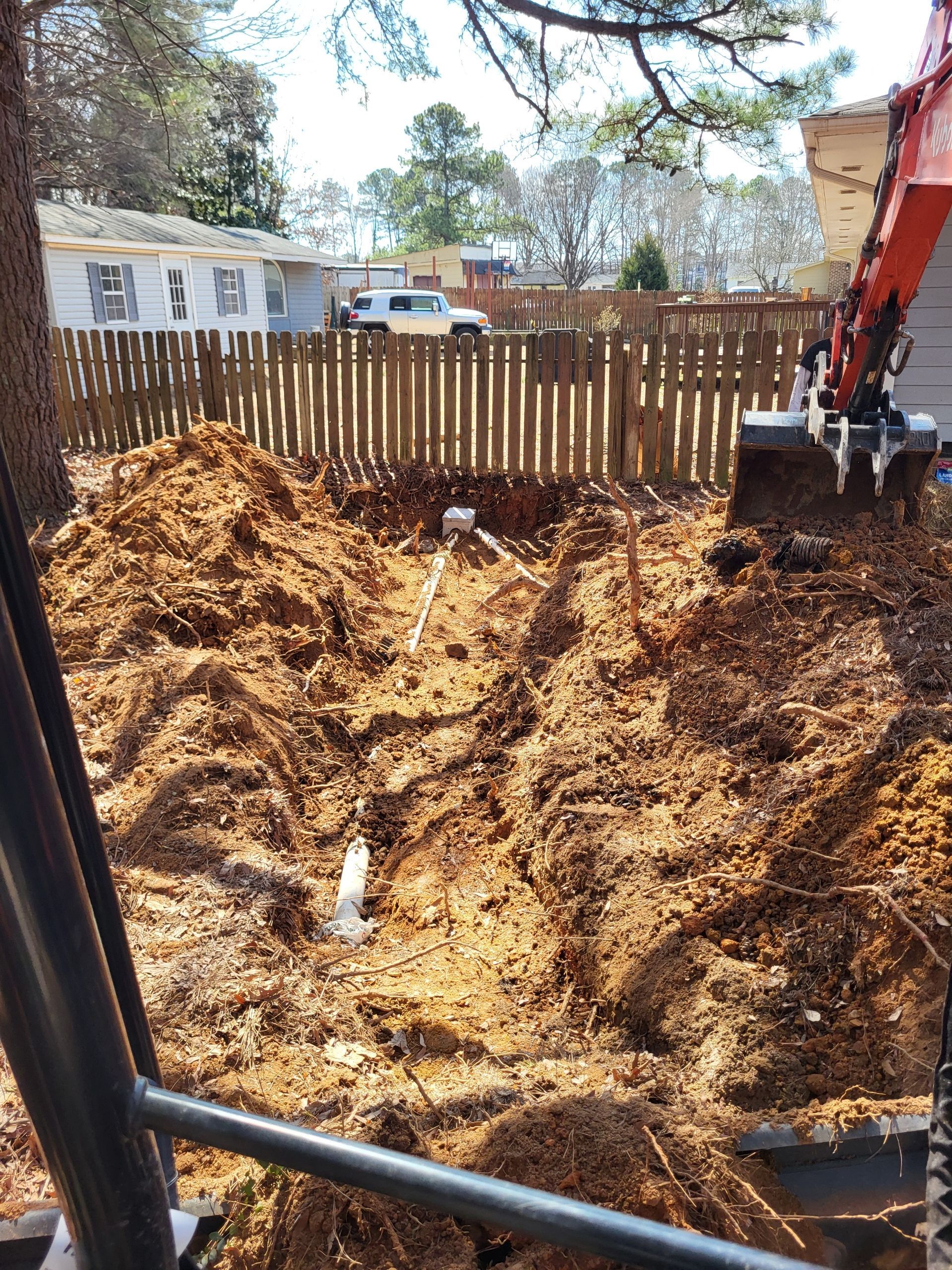 Excavator digging a large dirt pit beside a wooden fence in a backyard.
