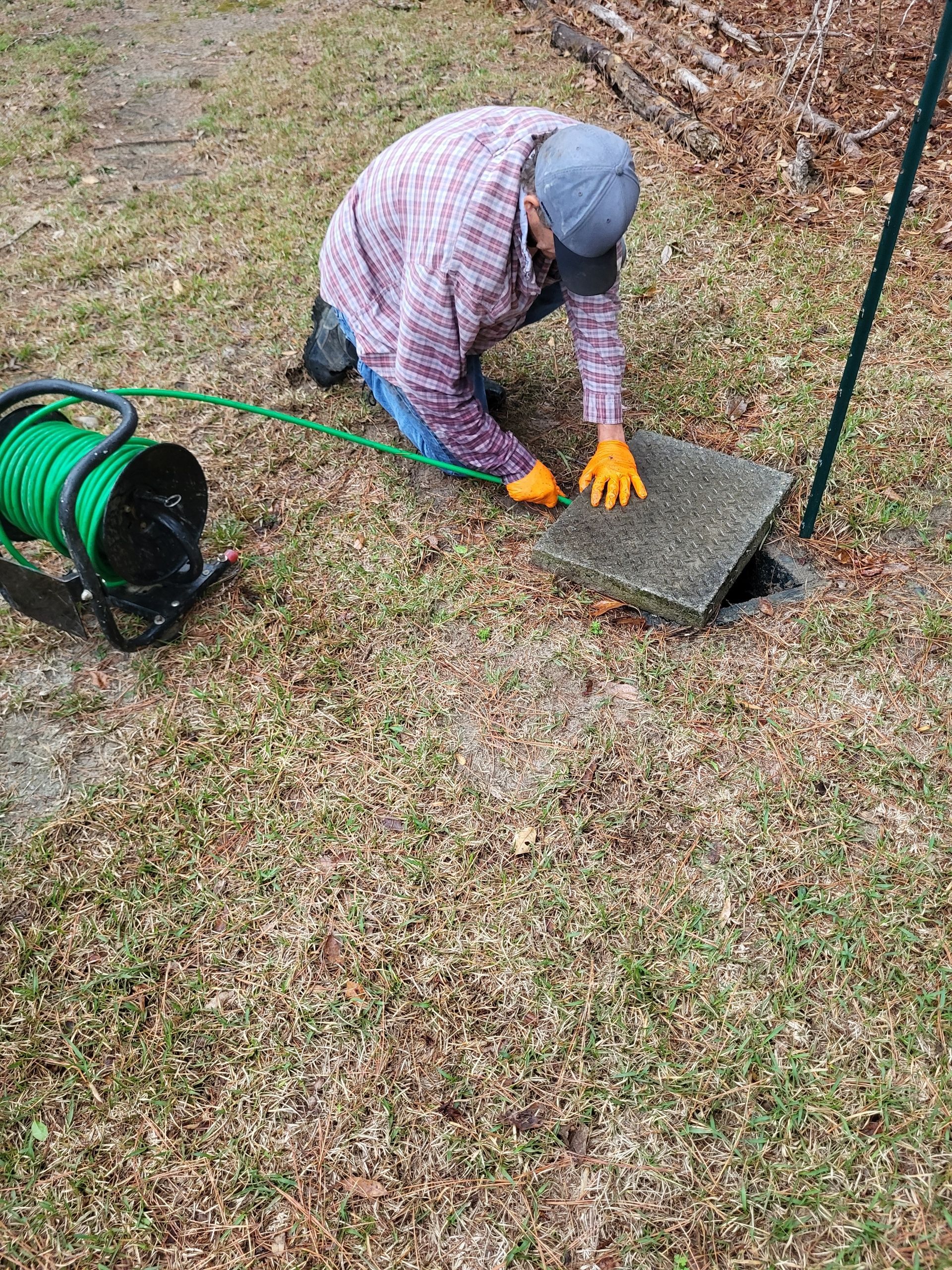 Person in gray cap and plaid shirt works on a concrete slab beside a green hose reel outdoors.