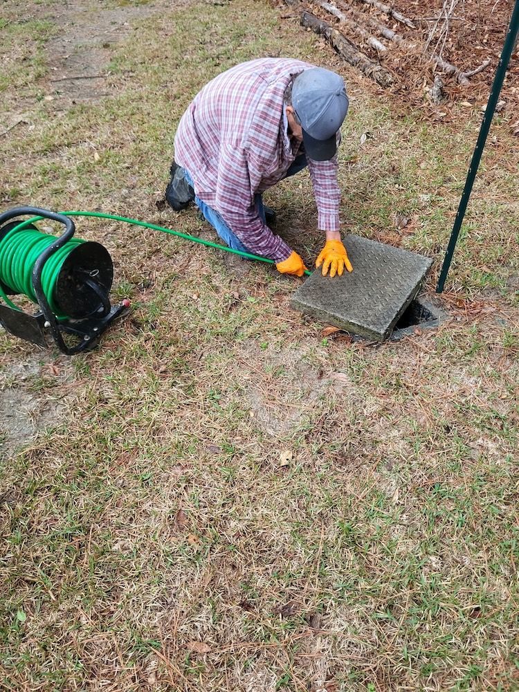 Person kneeling on grass cleaning a metal drain cover with orange gloves beside a green hose reel.