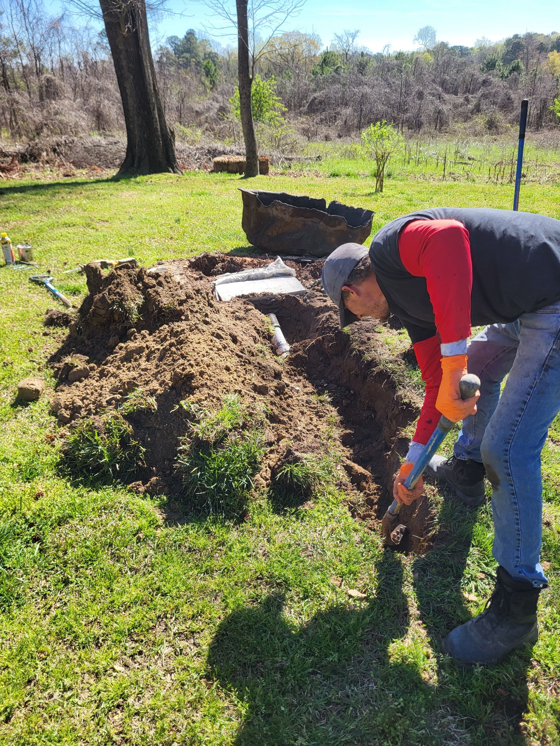Person digging in a garden beside a large pile of soil on a sunny grassy lawn