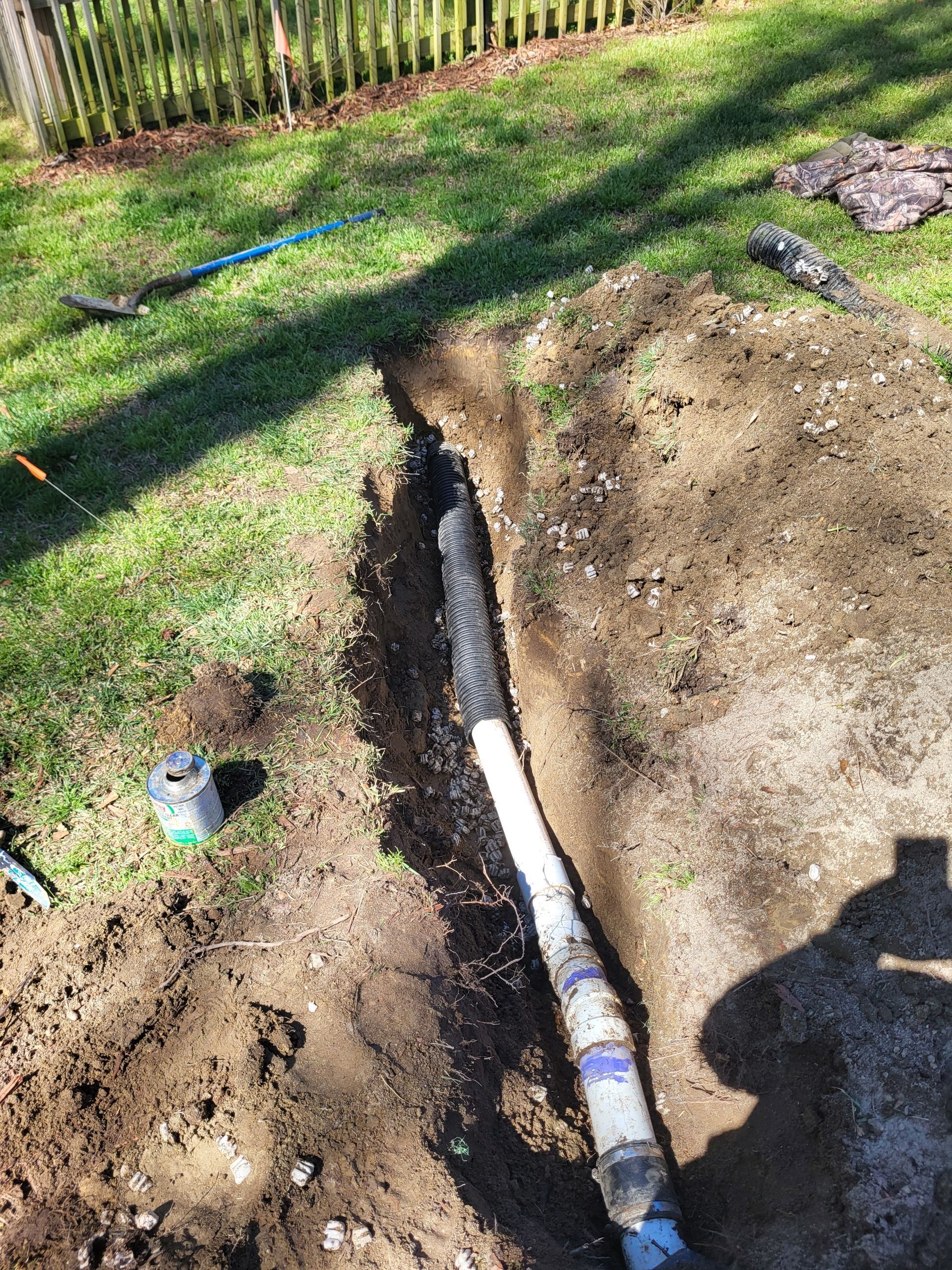 Trenched yard with a white pipe exposed in the ground beside a shovel and scattered dirt
