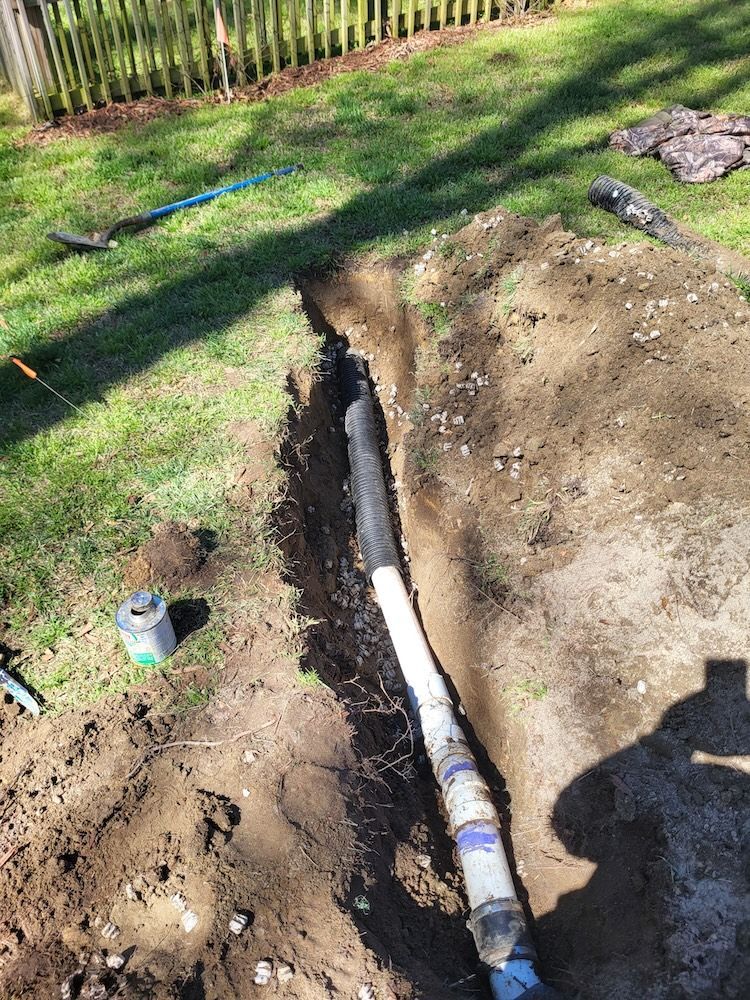 Trench in a dirt yard exposing a white pipe, with tools nearby and a fence in the background