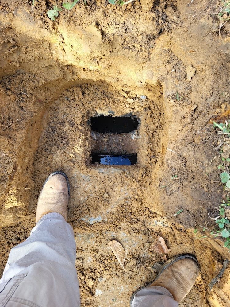 Boots beside a muddy excavation hole with a dark rectangular opening in the ground.