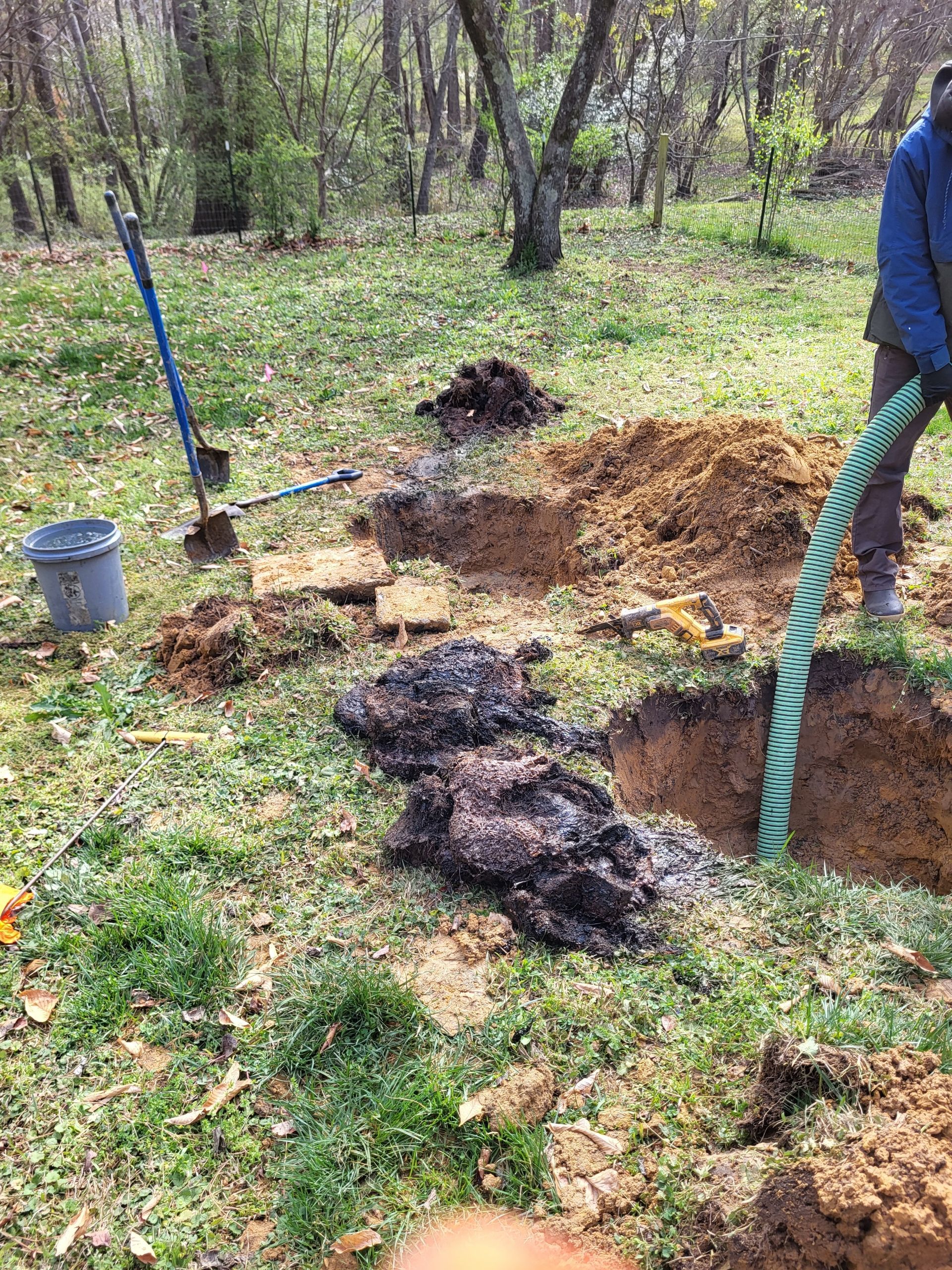 Person digging a large hole beside piles of soil in a grassy wooded area