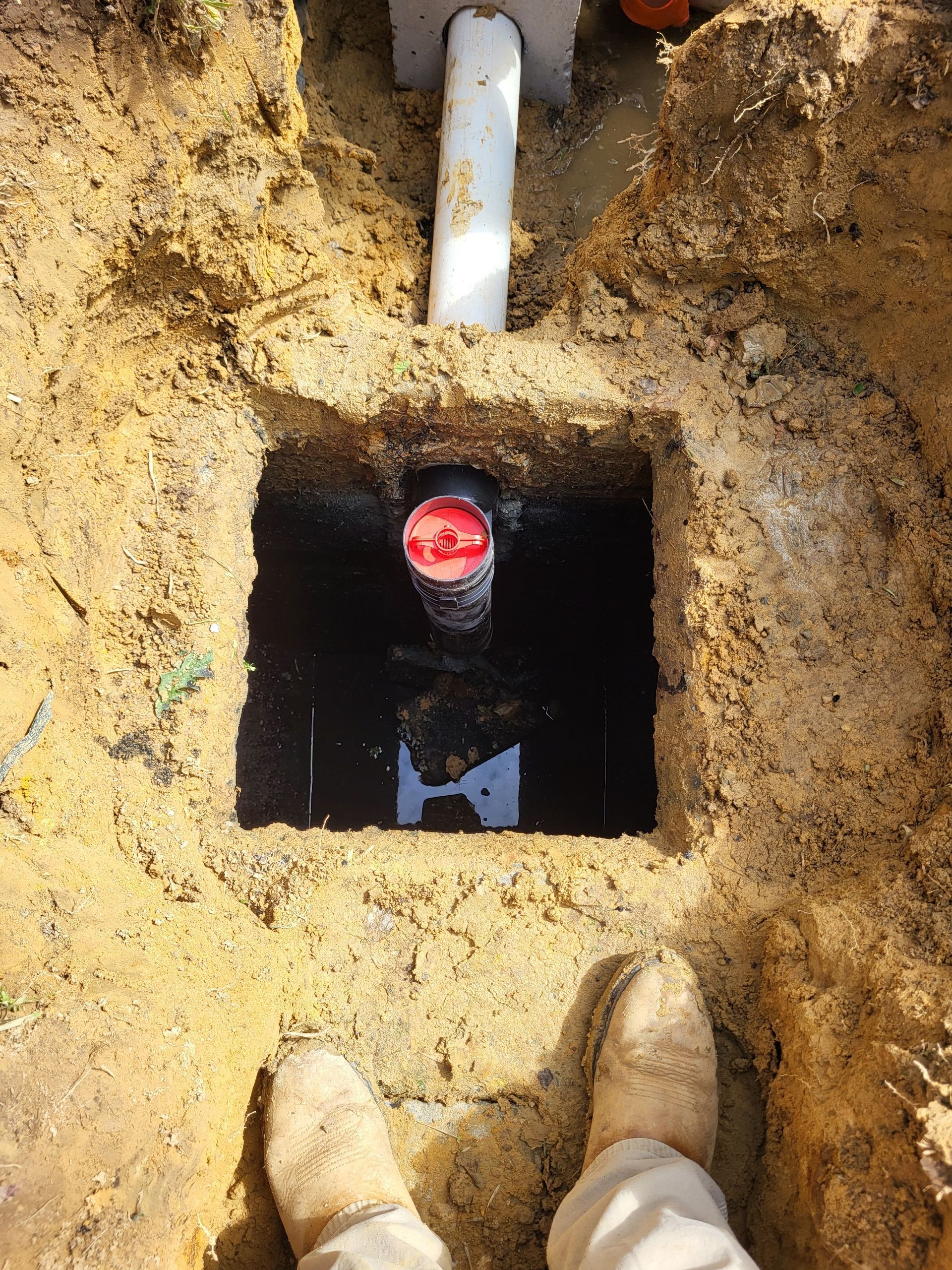 Open utility pit in dirt with a white pipe and red-capped valve, viewed from above with boots at the edge